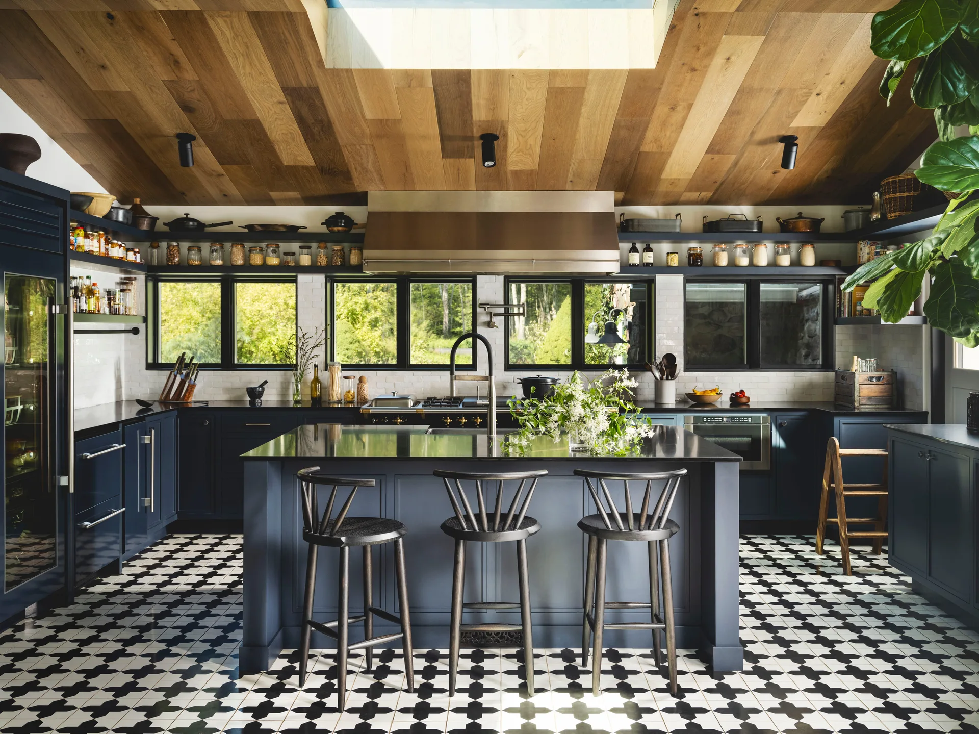 A residential kitchen features navy blue cabinetry, a geometric tiled floor, a central island with black wooden stools, and a vaulted wood plank ceiling.