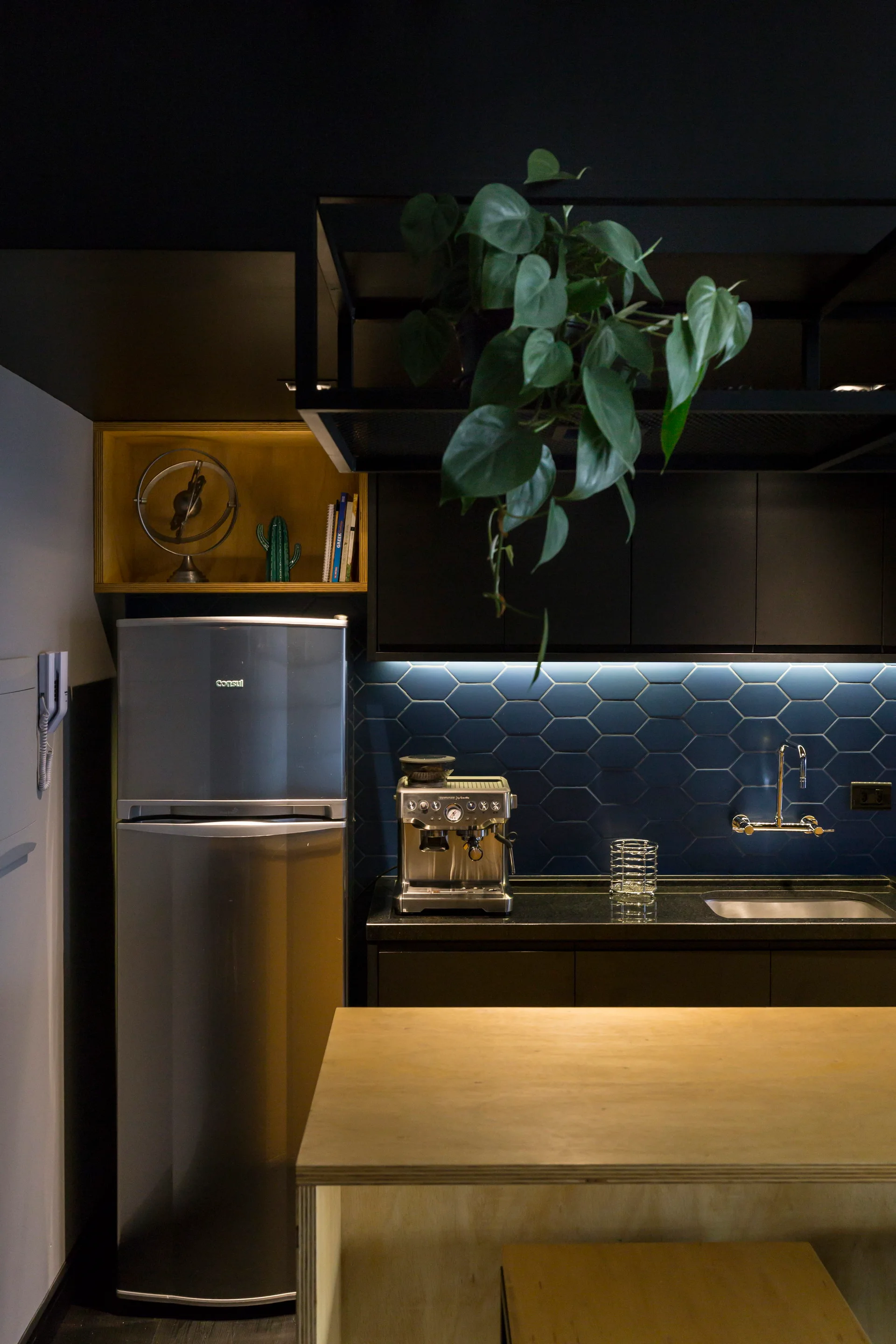 A dark modern kitchen features navy blue hexagonal tiles on the backsplash, matte black upper cabinets, and a light wood counter in the foreground.