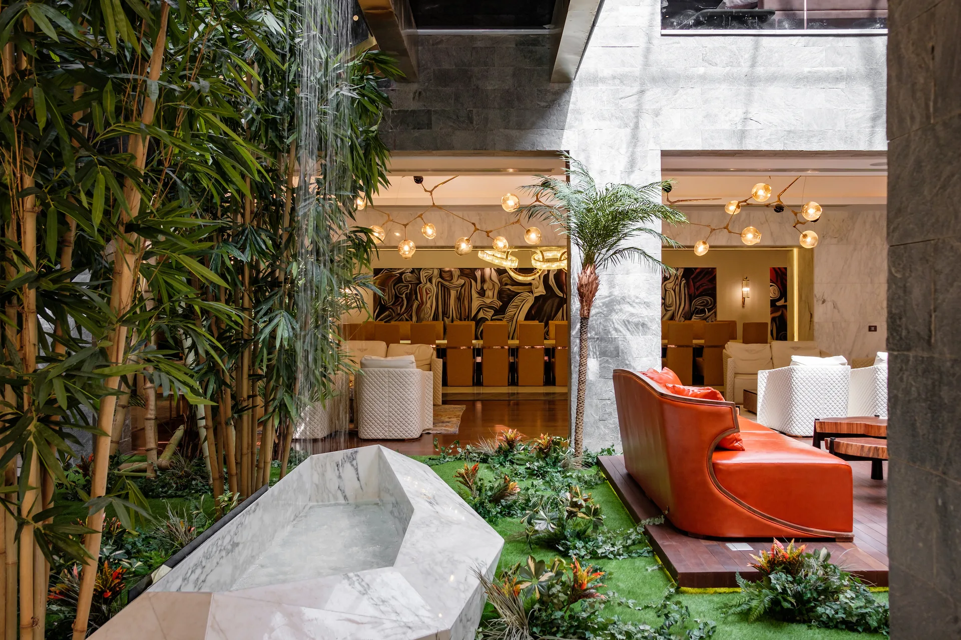 An indoor atrium featuring a white marble fountain with a waterfall, a curved orange leather sofa, stone walls, and bamboo plants.