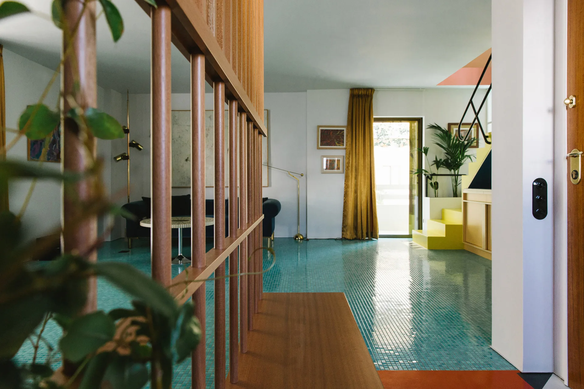 A living room features reflective turquoise mosaic floor tiles, a vertical wood slatted room divider, and bright yellow steps in the background.