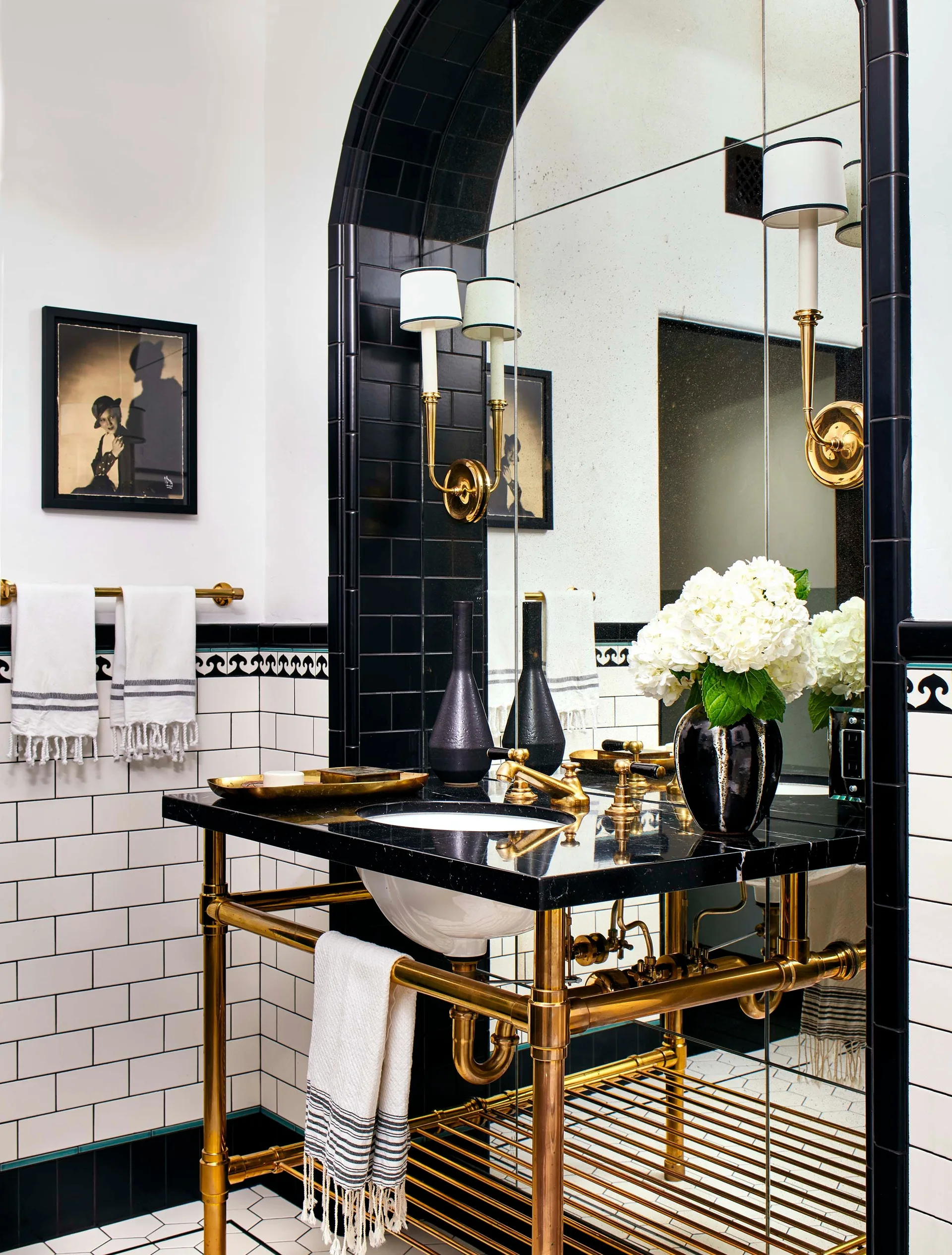 A bathroom featuring a black stone vanity with a polished brass frame set against white subway tiles and a large arched mirror framed in black tiles.