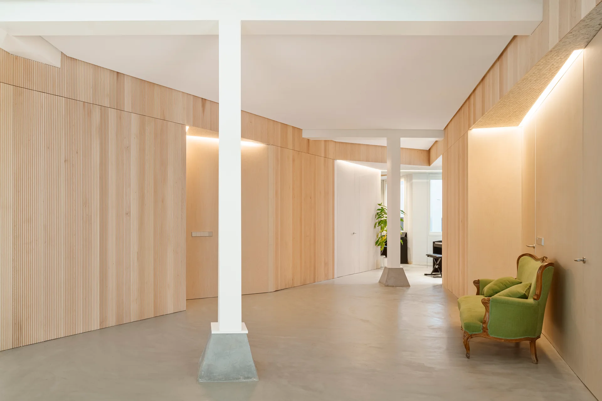 A minimalist hallway features ribbed light wood walls, concrete flooring, structural columns, and a vintage green armchair.