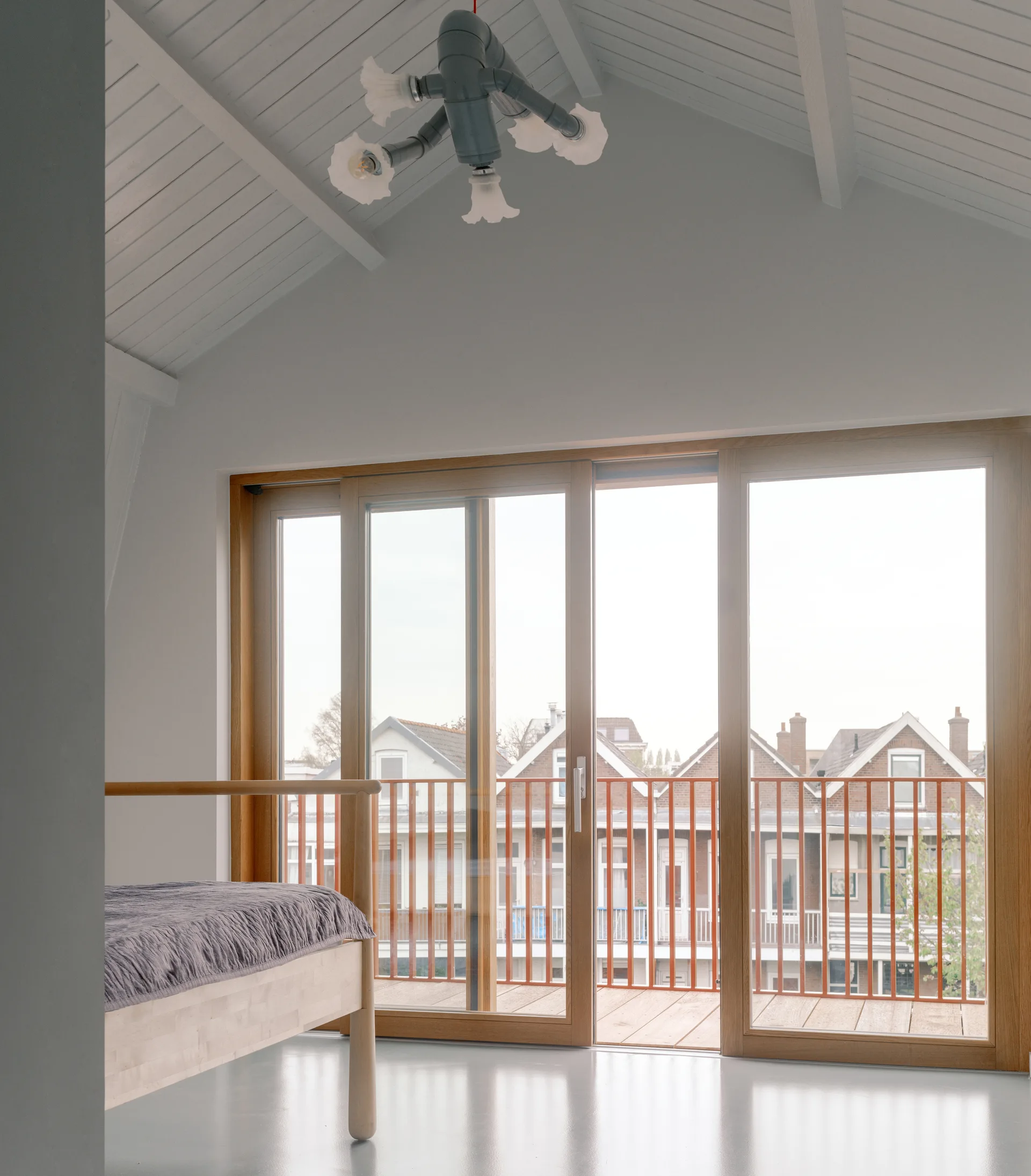 A minimalist bedroom features a sloped white wood ceiling with a pipe chandelier, a wooden bed, and sliding glass doors opening to a balcony.