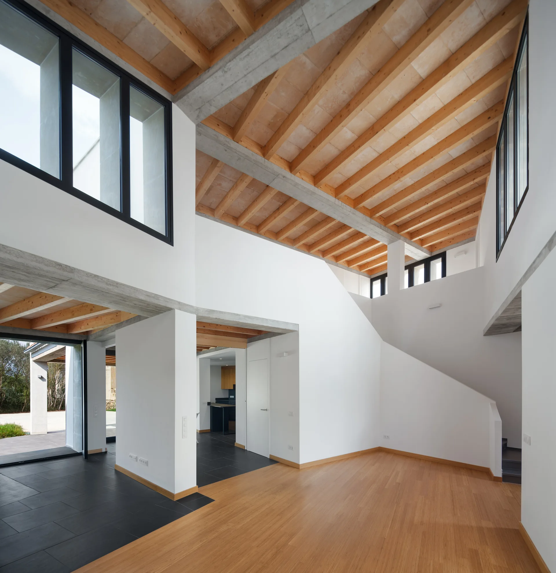 A modern double-height living room features white walls, exposed wooden ceiling beams with concrete supports, and a floor divided between wood and dark tile.