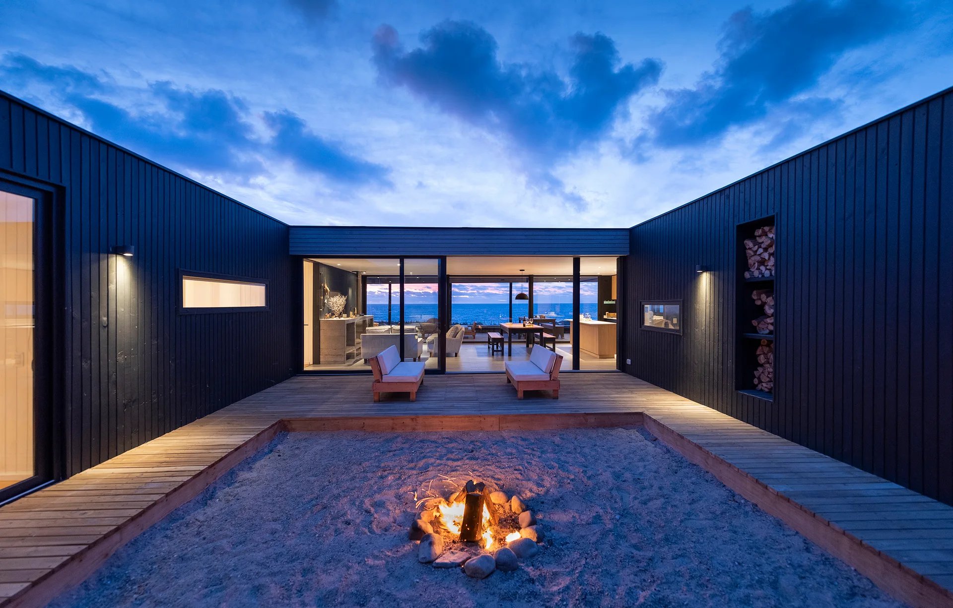 A modern courtyard features a central stone fire pit in a sandy area surrounded by a wooden deck and dark charcoal siding at twilight.