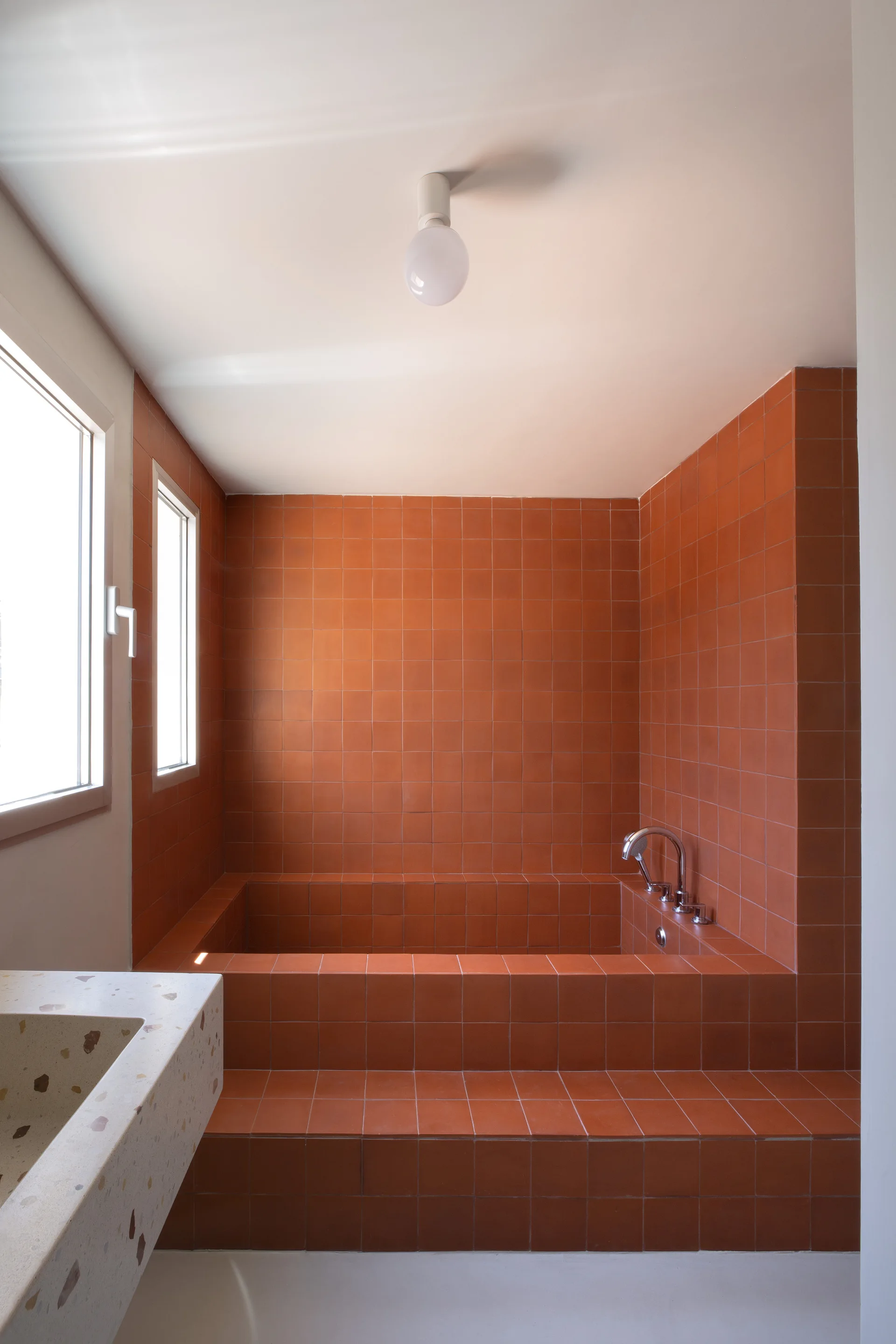 A minimalist bathroom features a built-in bathtub and walls clad in terracotta square tiles alongside a speckled terrazzo vanity.
