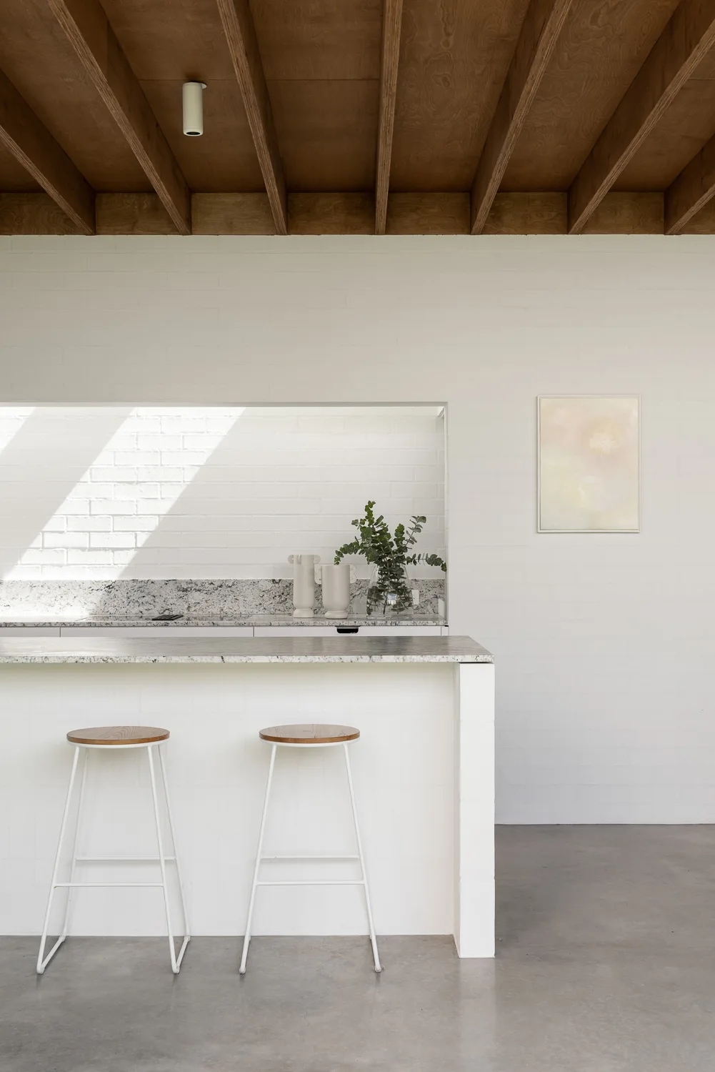 A minimalist kitchen featuring a white brick wall, an island with two wood-topped stools, and exposed wooden ceiling beams.