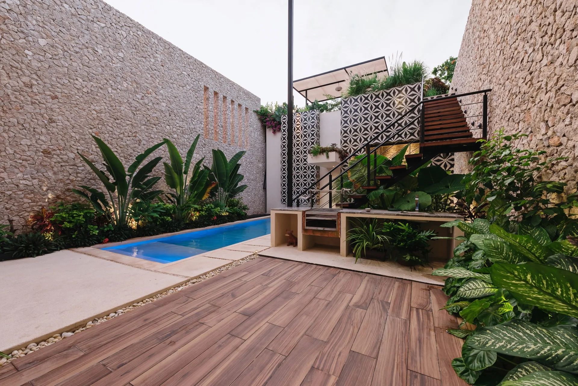 A narrow lap pool sits in an outdoor courtyard flanked by textured stone walls, a wooden deck, and a floating staircase with geometric wall tiles.