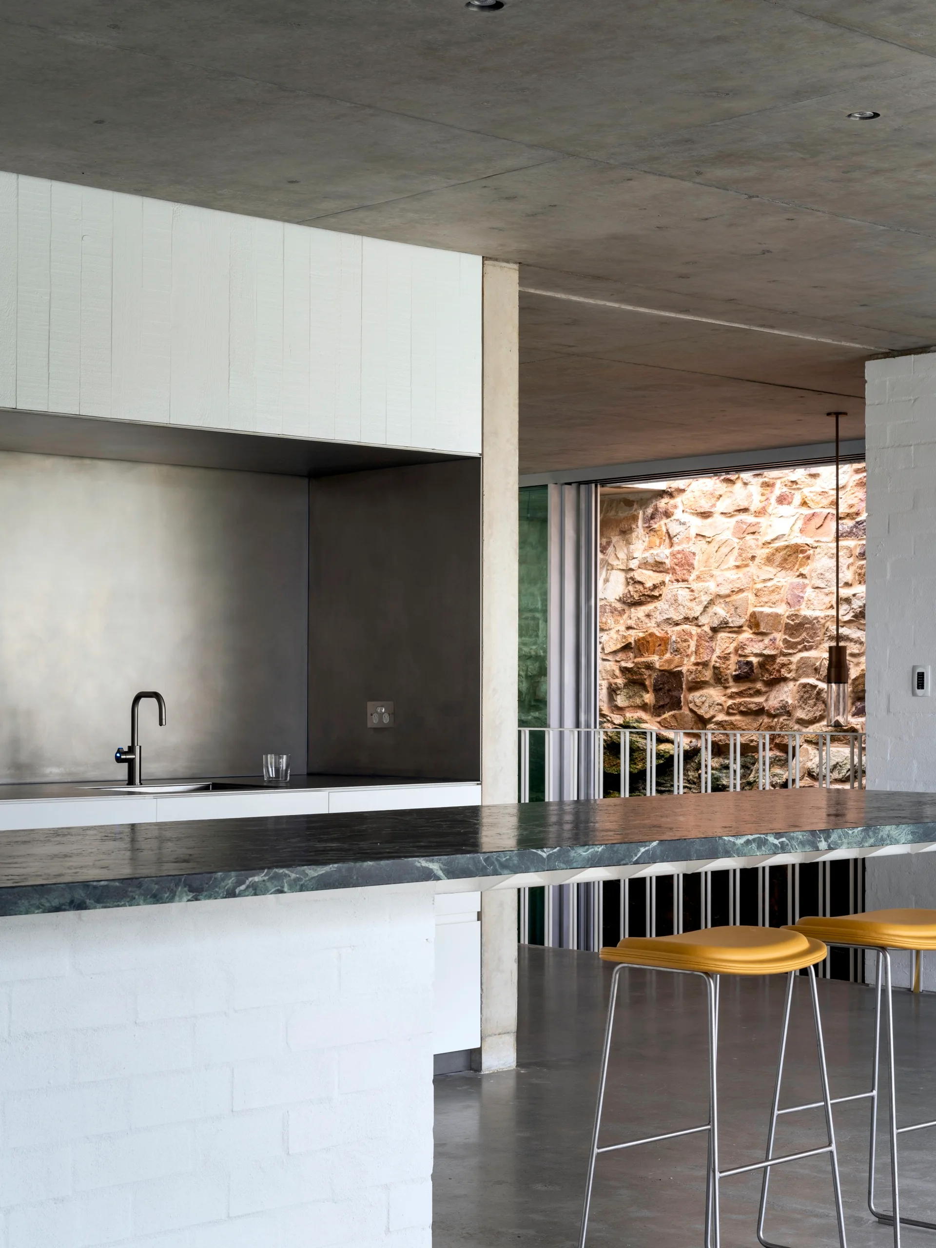 A modern kitchen features a green marble island with yellow stools under a concrete ceiling, facing a metallic backsplash and stainless steel sink.