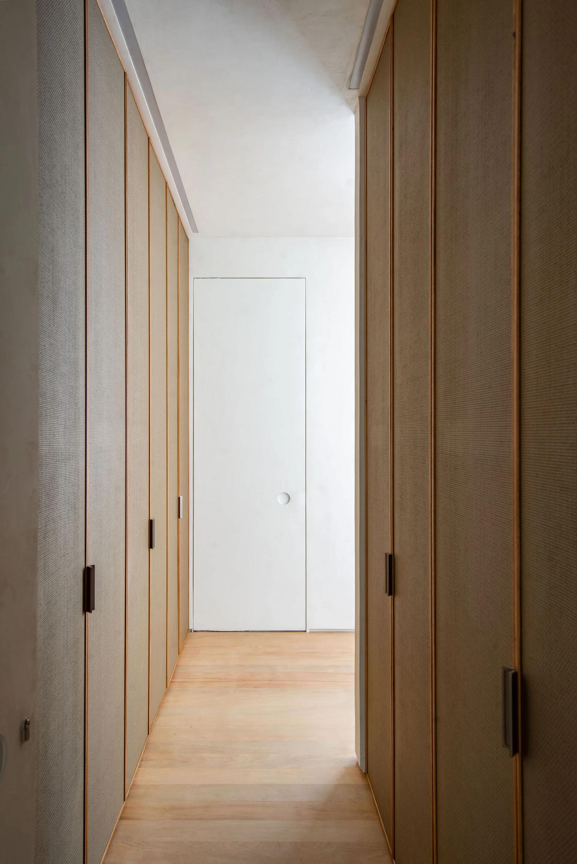 A narrow hallway with light wood flooring features floor-to-ceiling textured beige built-in wardrobes leading to a flush white door at the end.