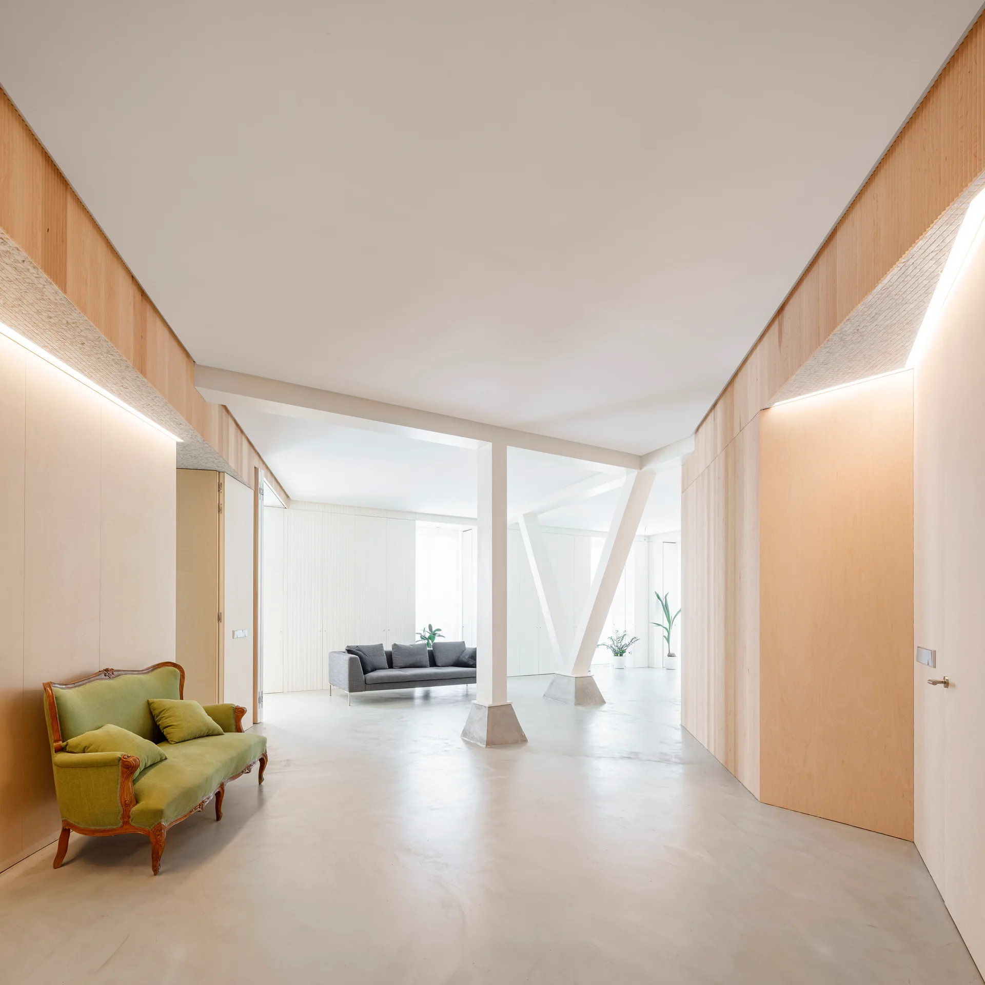 A minimalist open-plan interior features light wood-paneled walls, a green vintage settee, white support columns, and a grey sofa in the background.