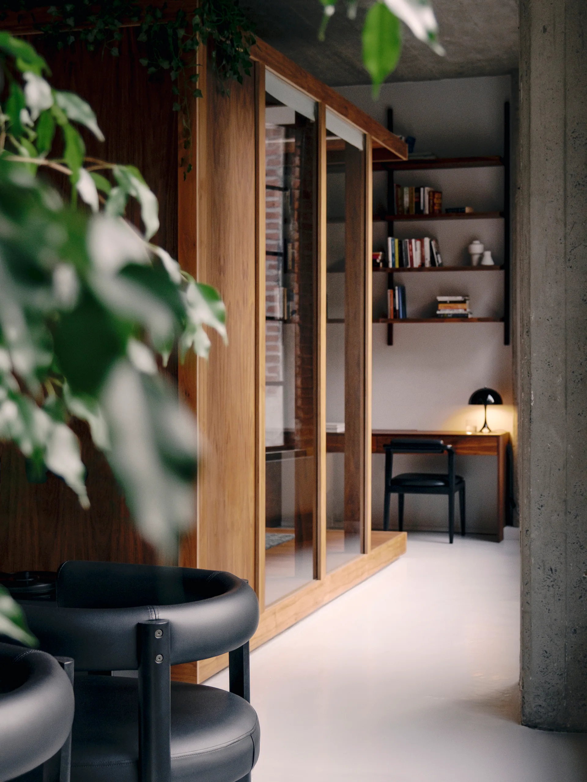 A modern home office features a wooden desk, a black leather chair, wall-mounted bookshelves, a concrete column, and a large wood-and-glass partition.