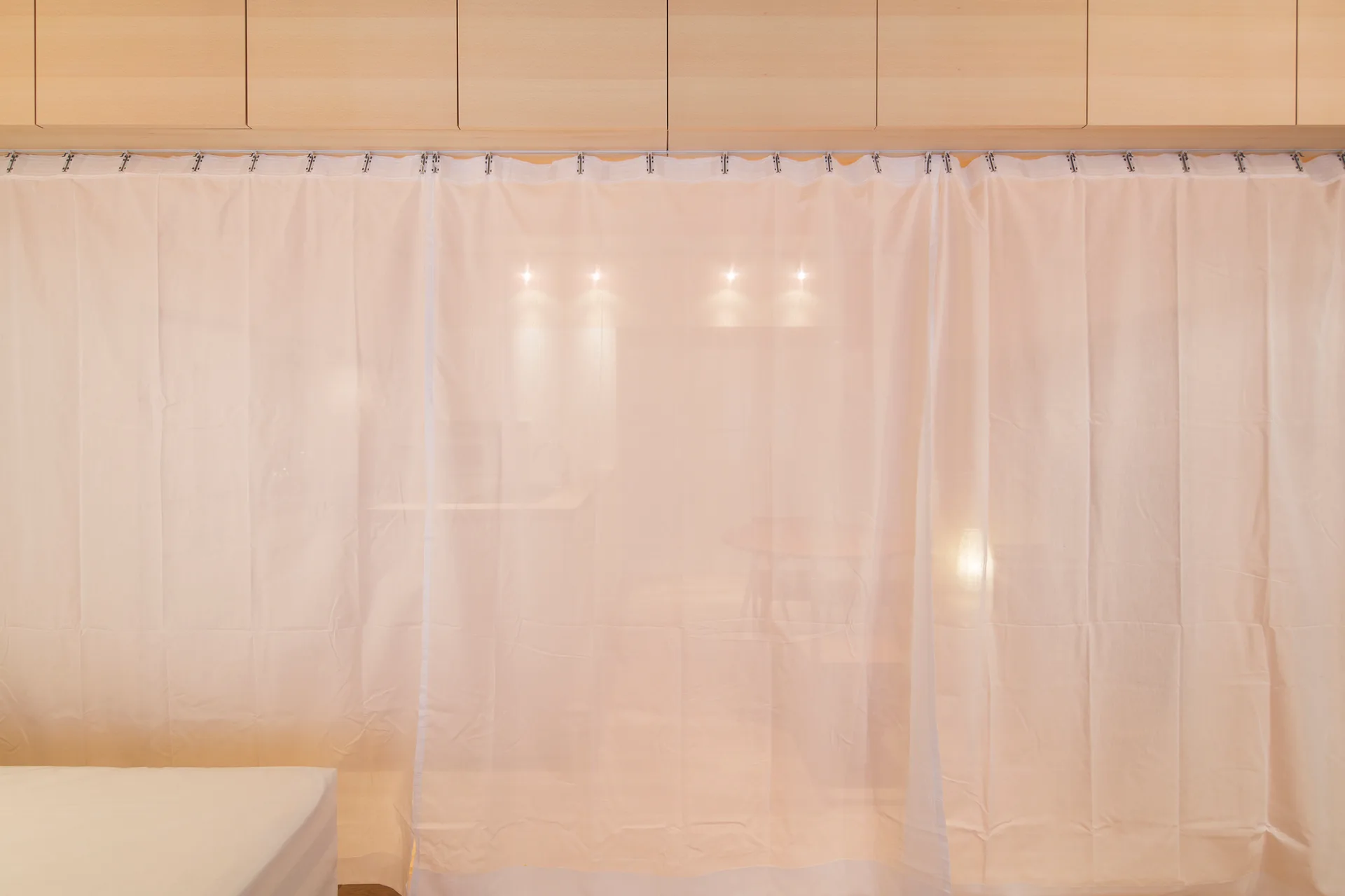 A sheer white curtain hangs from a track beneath light wood cabinetry, partially obscuring a warmly lit living area.