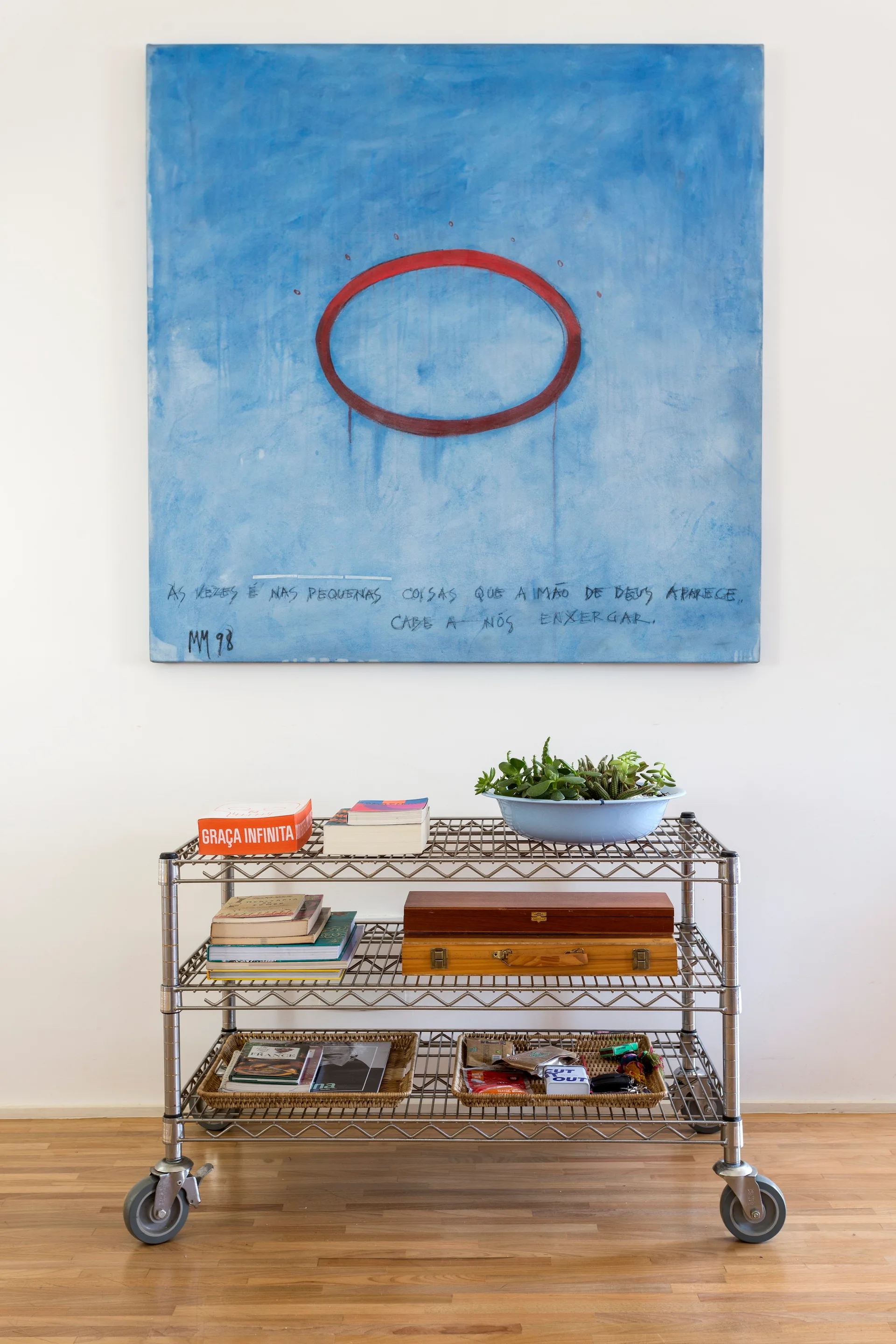 A chrome wire utility cart styled with books and succulents sits beneath a large abstract blue painting on a white wall.