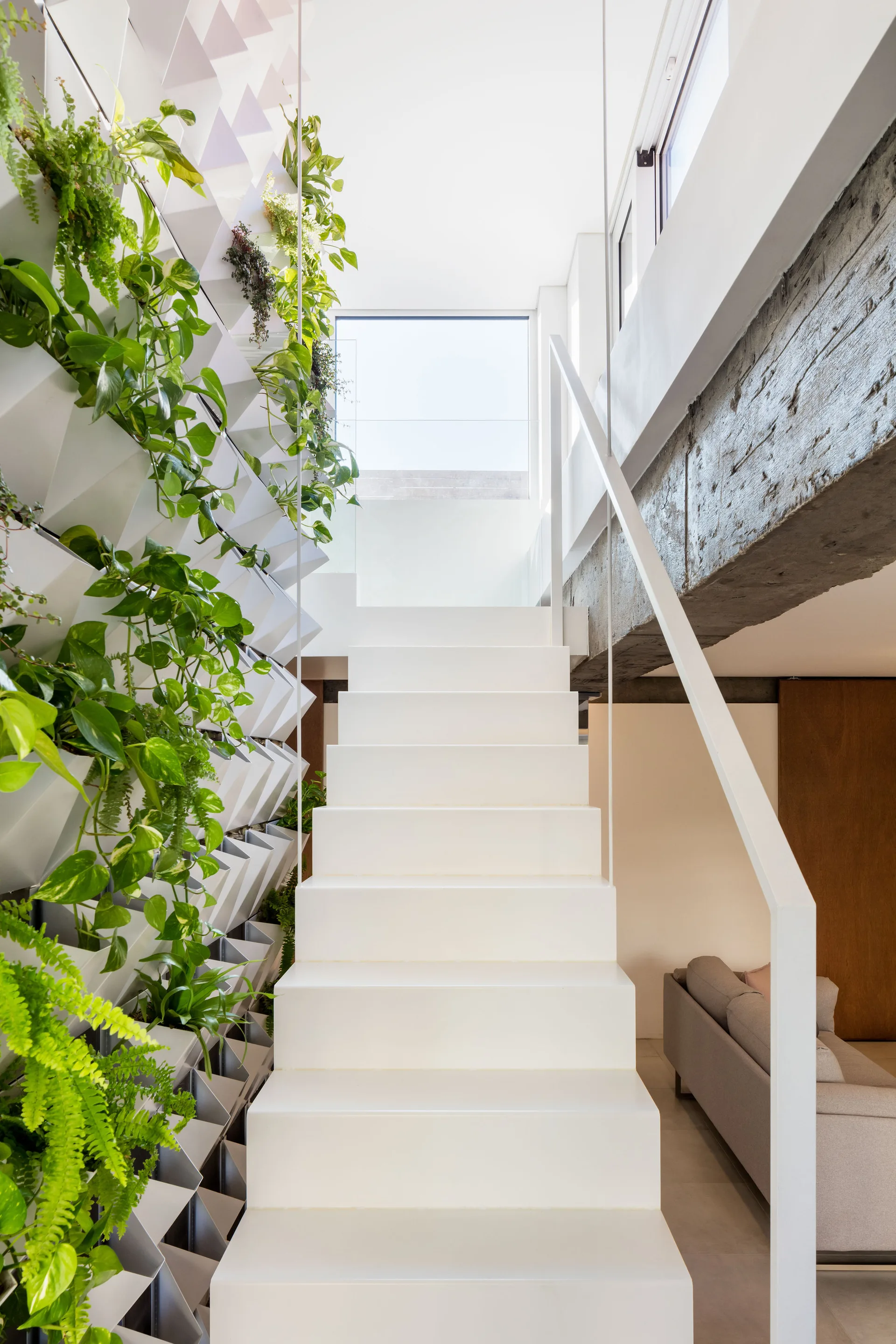 A modern white staircase is flanked by a geometric vertical living wall of lush green plants and an exposed concrete beam.
