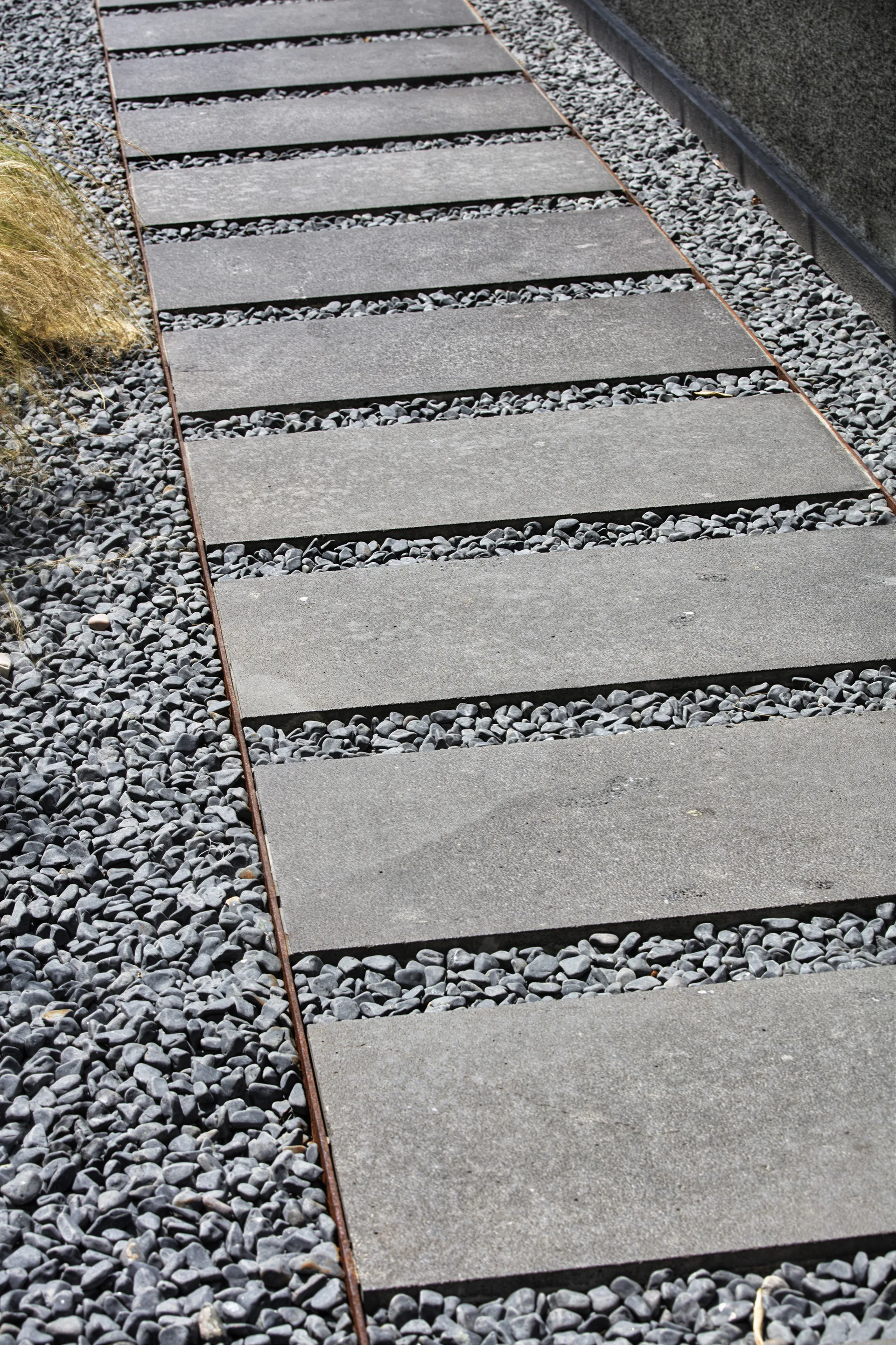 A garden path features large rectangular grey stone pavers set between bands of small dark grey pebbles and bounded by thin metal edging.