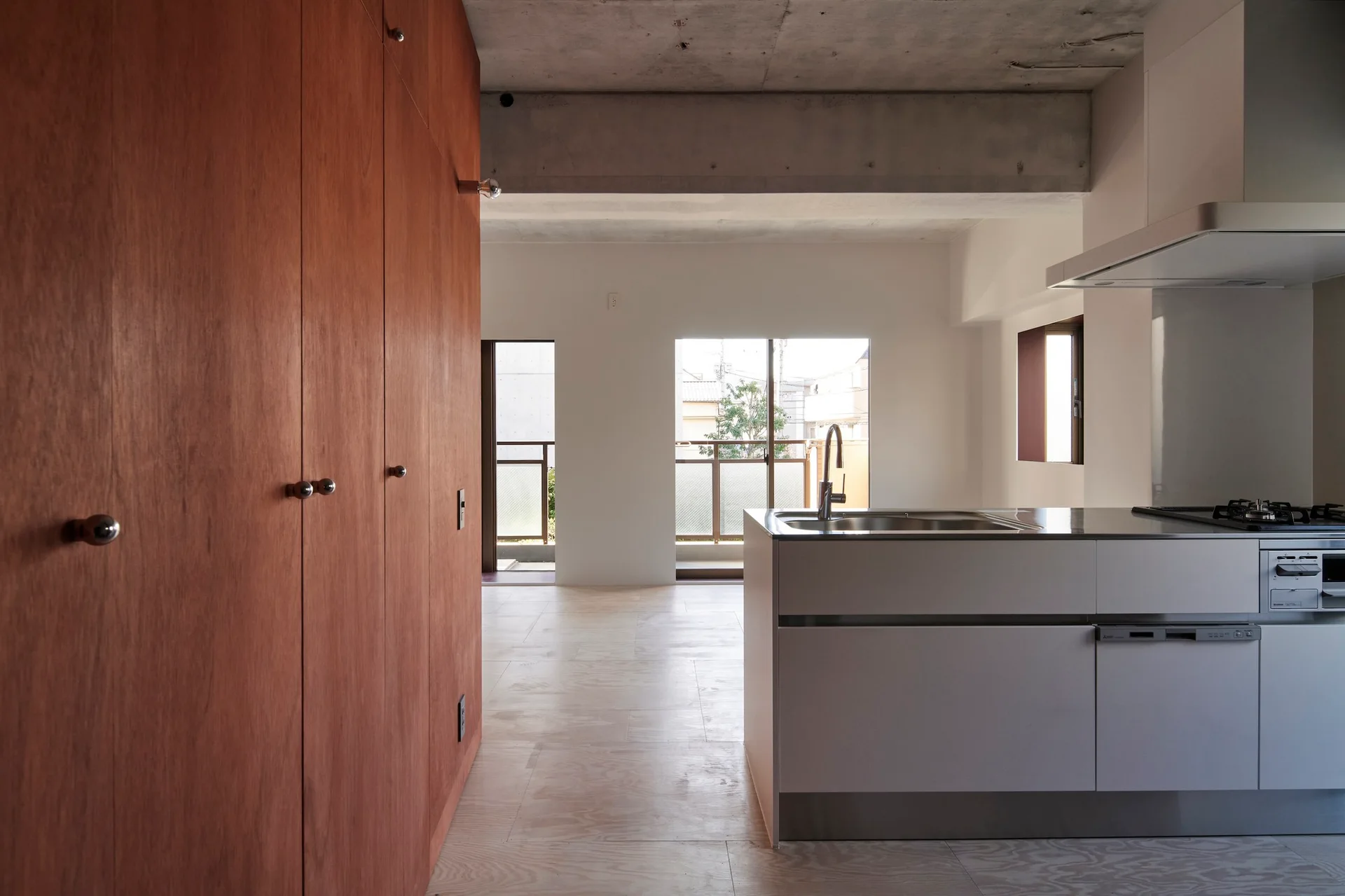 A minimalist kitchen features a tall wood storage wall, a modern white island with a sink, and an exposed concrete ceiling.