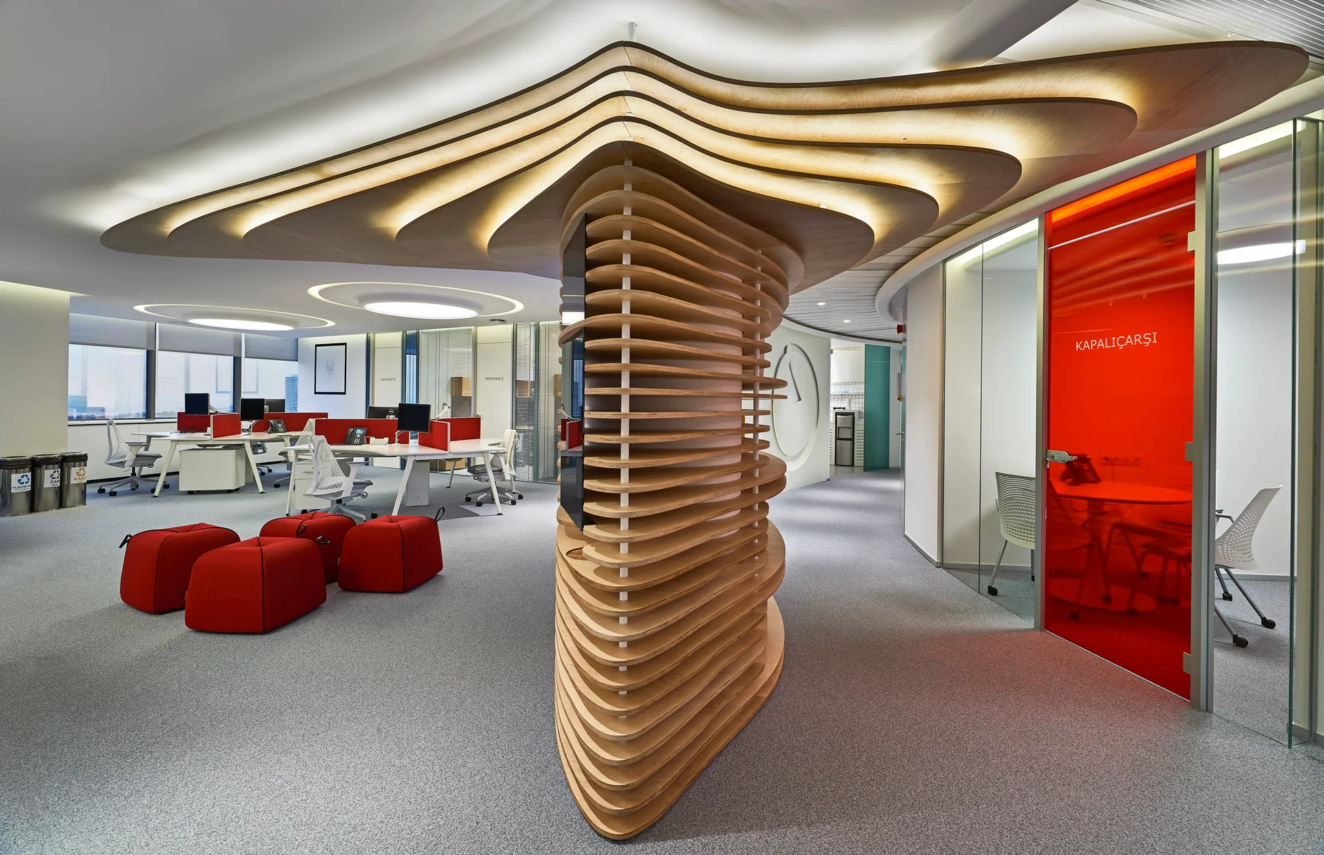 A modern open-plan office features a central layered-wood sculptural partition beneath integrated lighting, with red chairs and a glass office door nearby.