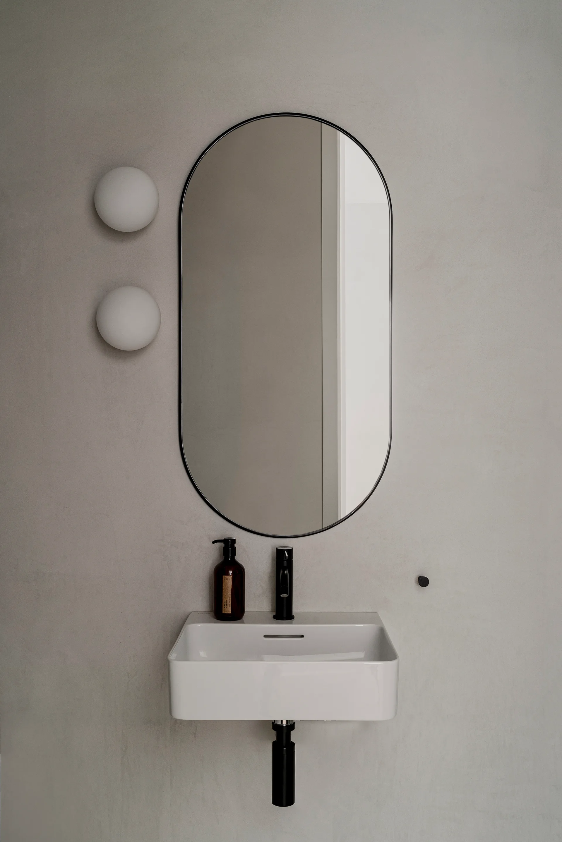 An oval black-framed mirror hangs above a white wall-mounted sink with two white globe sconces on a textured grey plaster wall.