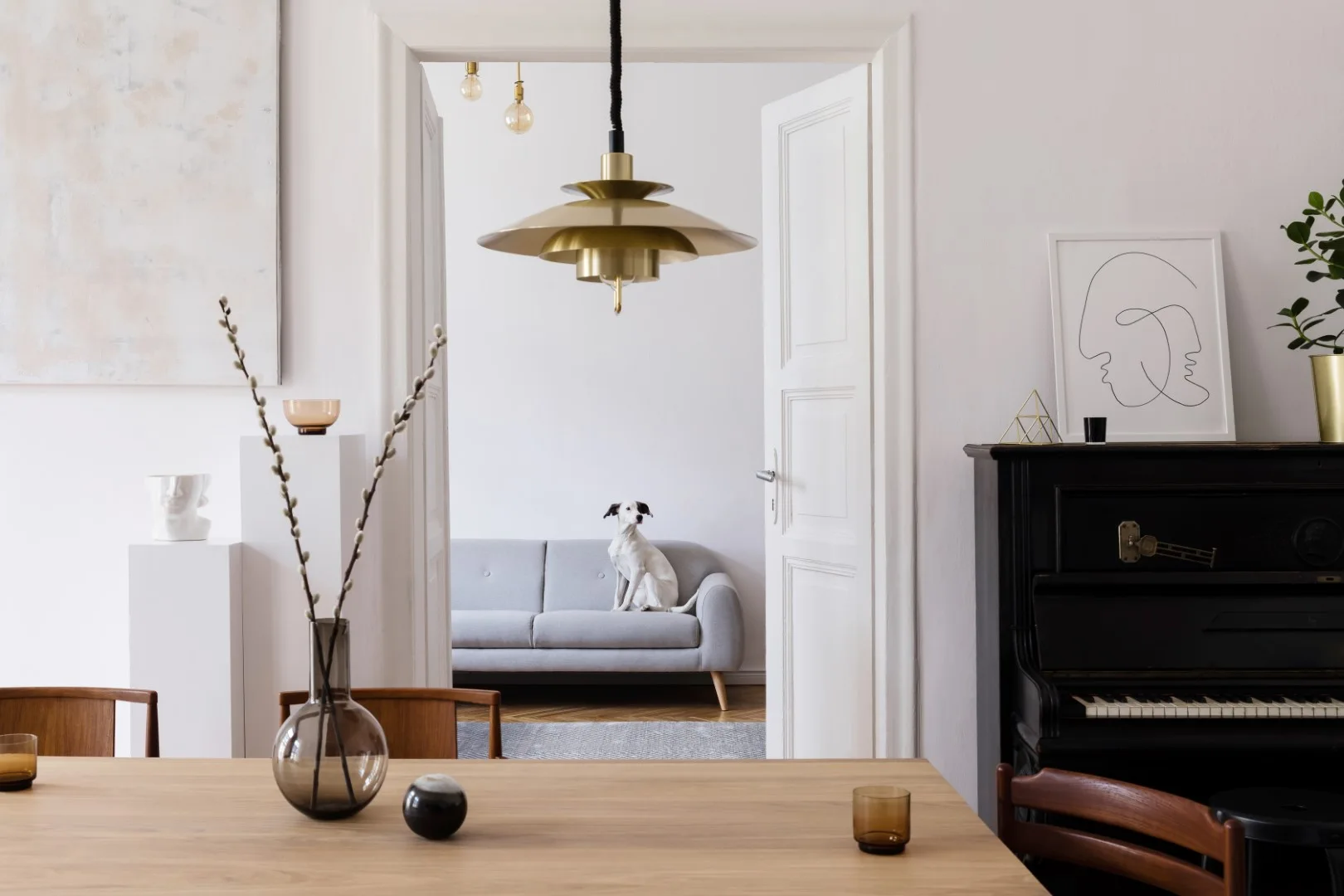A view from a wooden dining table through white double doors into a living room featuring a grey sofa, a brass pendant light, and a black piano.