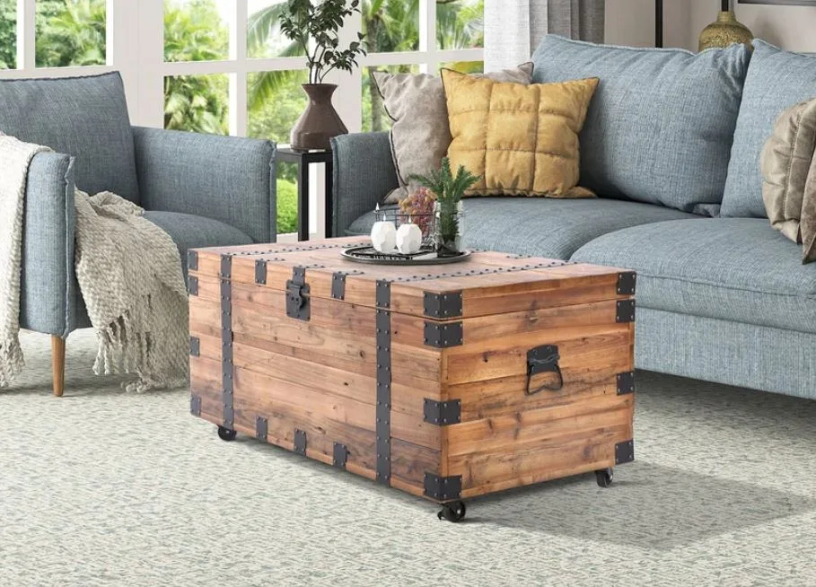 A wheeled wooden storage trunk with black corner brackets acts as a coffee table in front of a blue upholstered sofa.