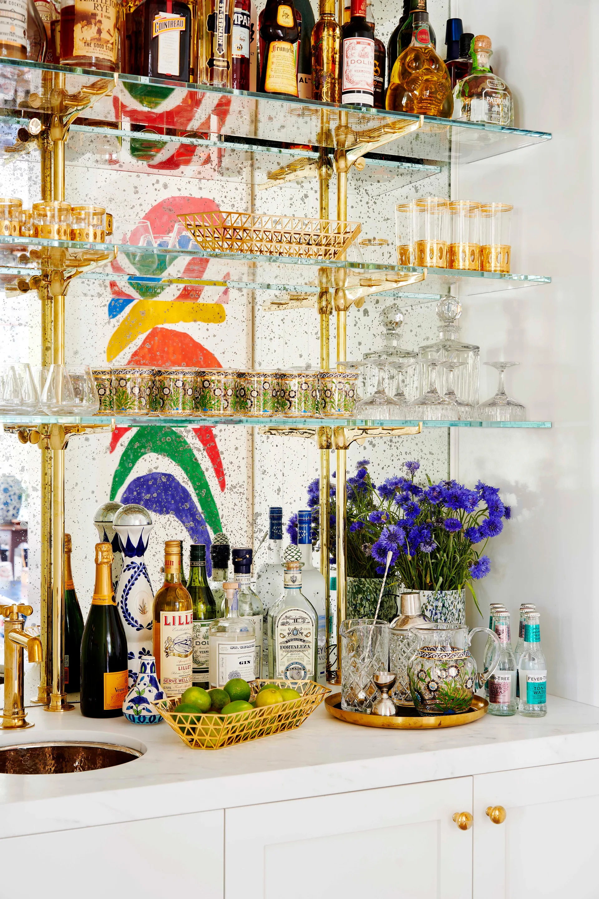 A home bar featuring brass and glass shelving against an antiqued mirror backsplash with various bottles and glassware.