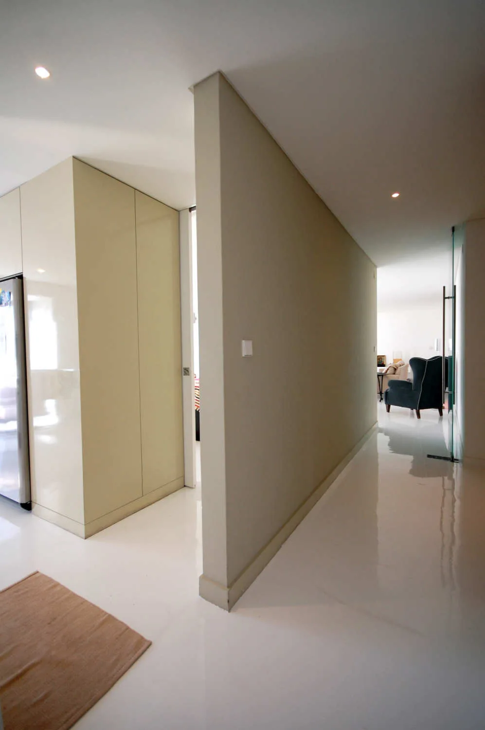 A minimalist residential hallway features highly reflective white flooring, cream built-in storage cabinets, and a view of a blue armchair in the distance.