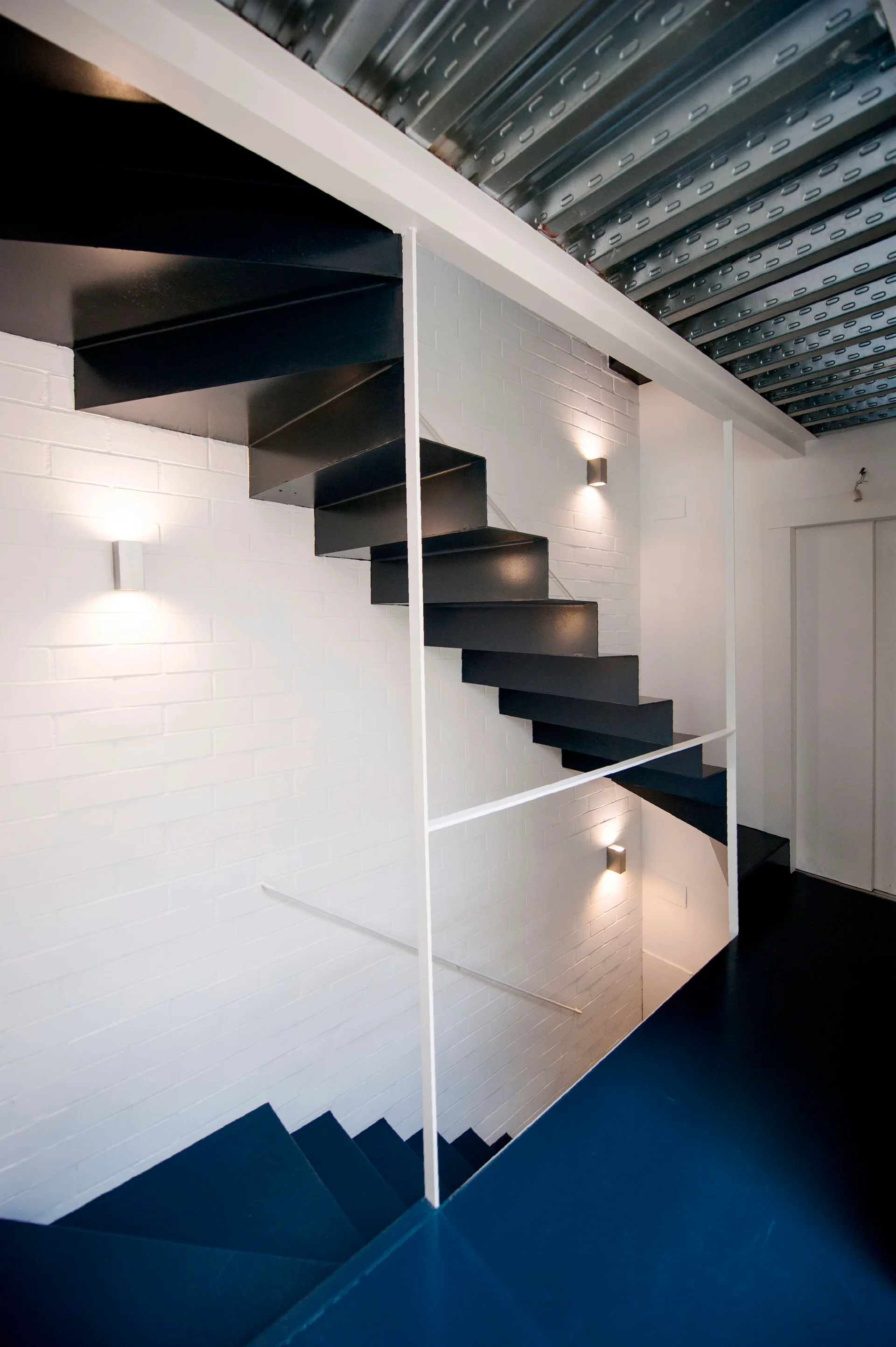 A modern floating black metal staircase stands against a white painted brick wall with wall sconces beneath an exposed corrugated metal ceiling.