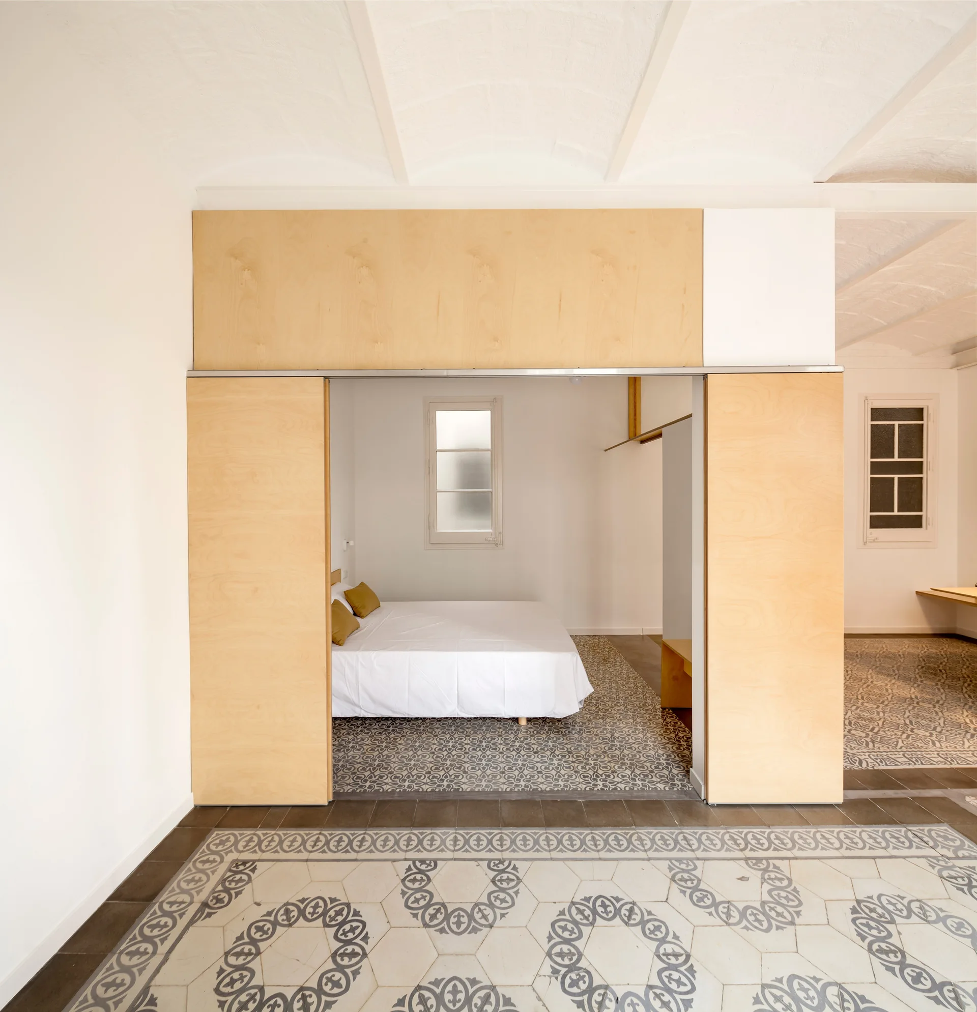 A minimalist bedroom featuring a bed with yellow pillows set behind a sliding light wood partition wall on patterned ceramic tile floors.