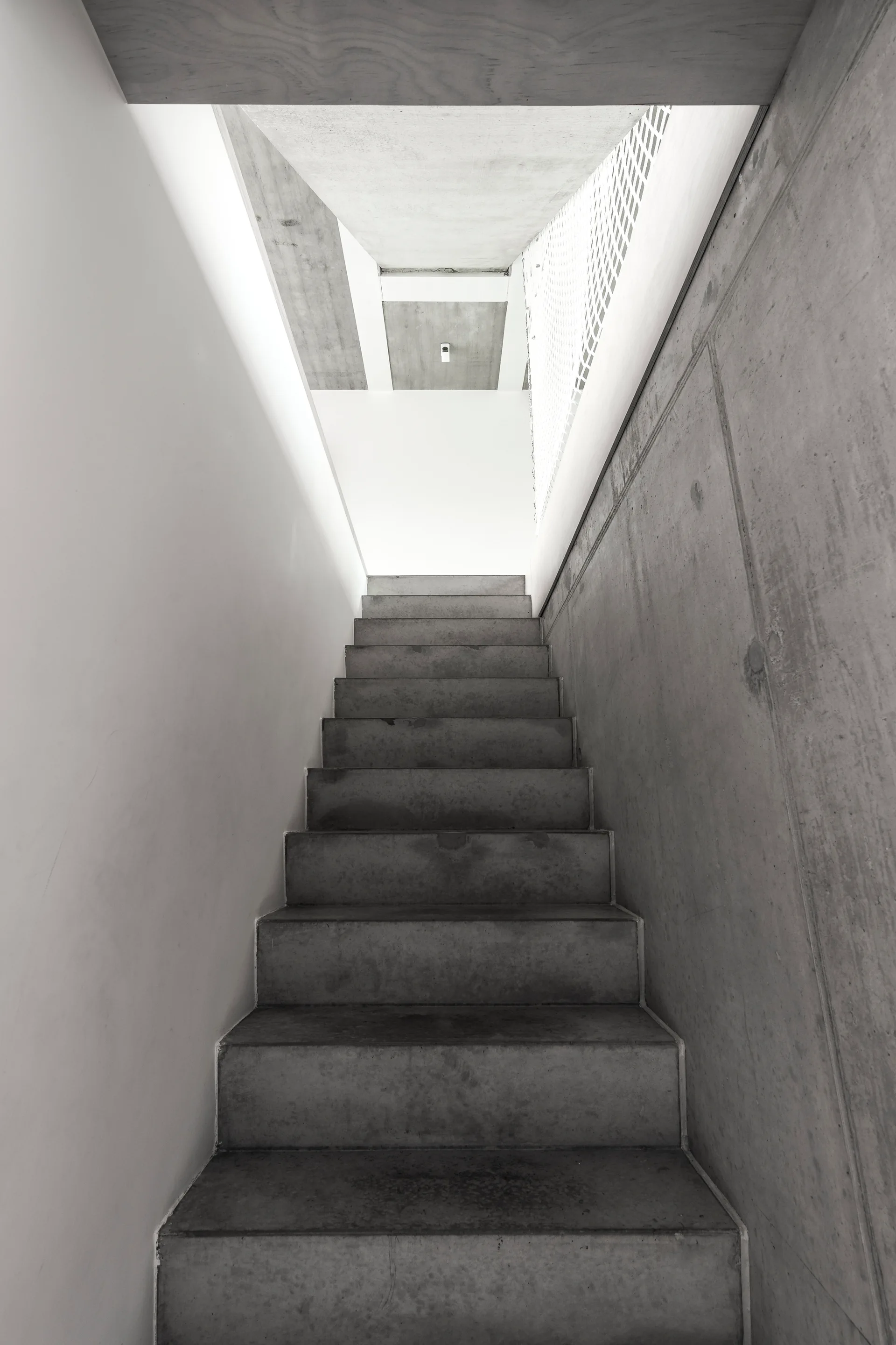 A narrow concrete staircase ascends between a smooth white wall and a textured board-formed concrete wall toward a bright landing.