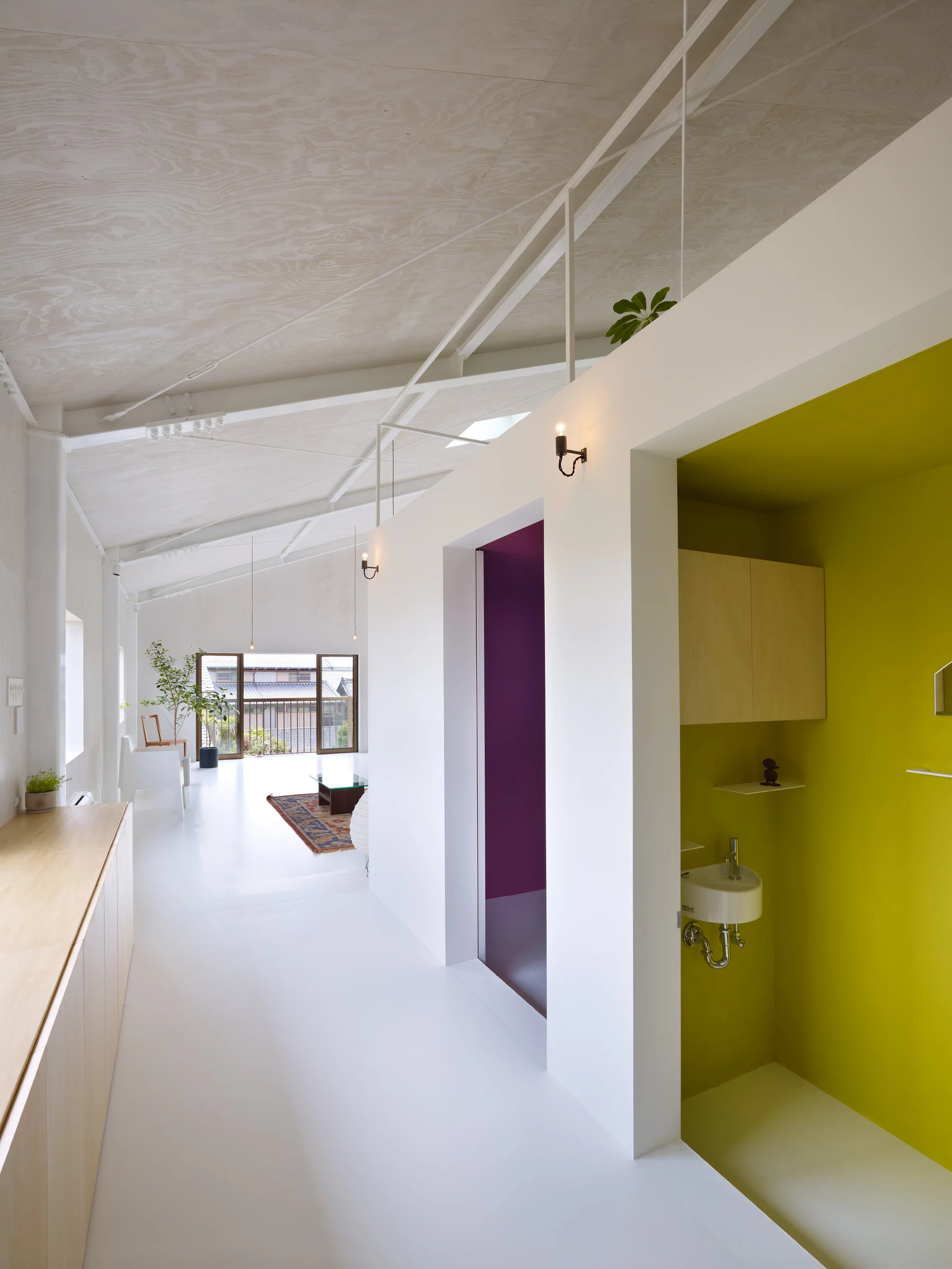 A long minimalist white hallway features a sloped plywood ceiling, a bright yellow alcove with a small sink, and built-in wood cabinets.