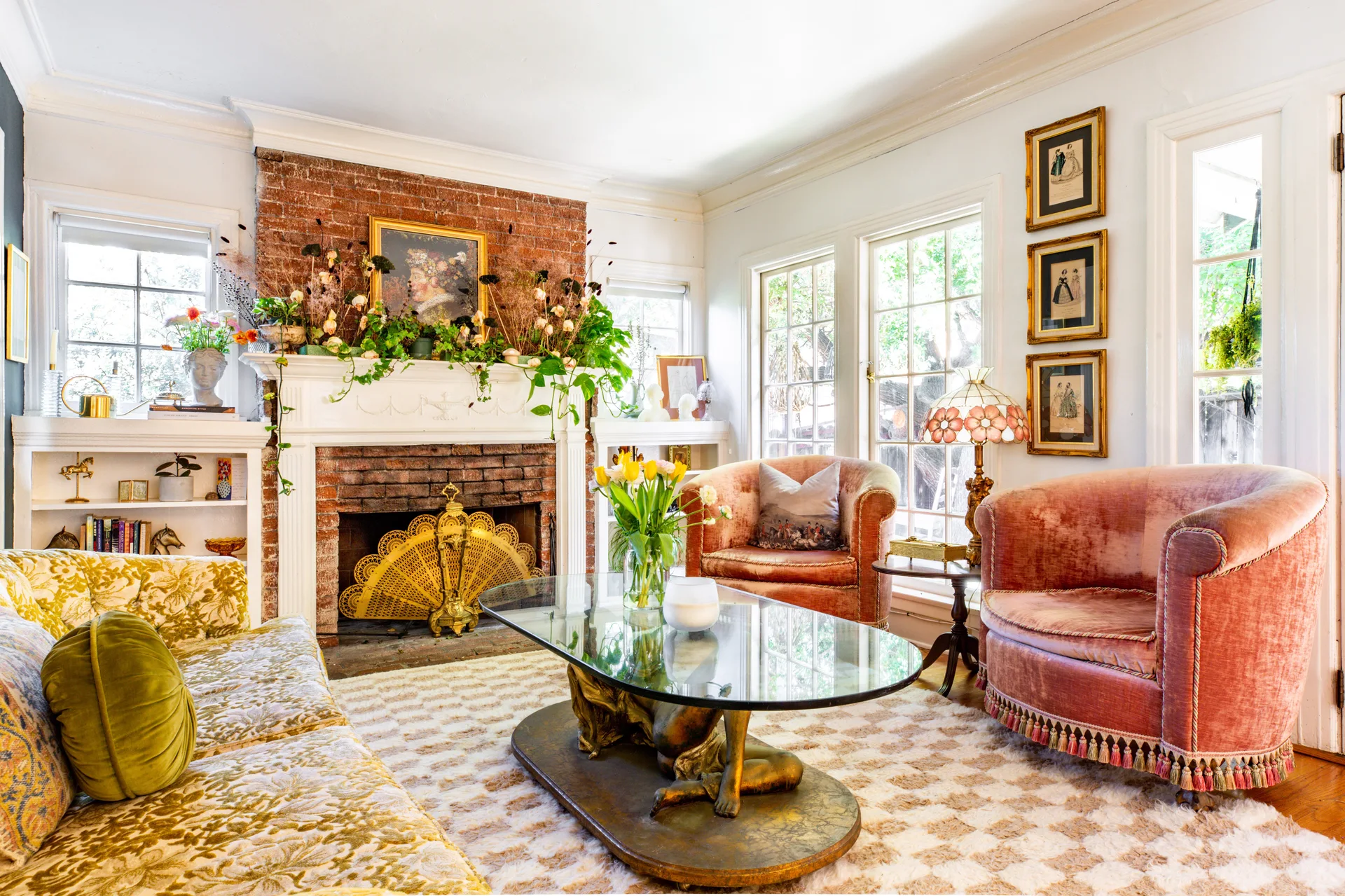 A maximalist living room features a brick fireplace with a white mantel, two pink velvet armchairs with tassel trim, and a glass coffee table.