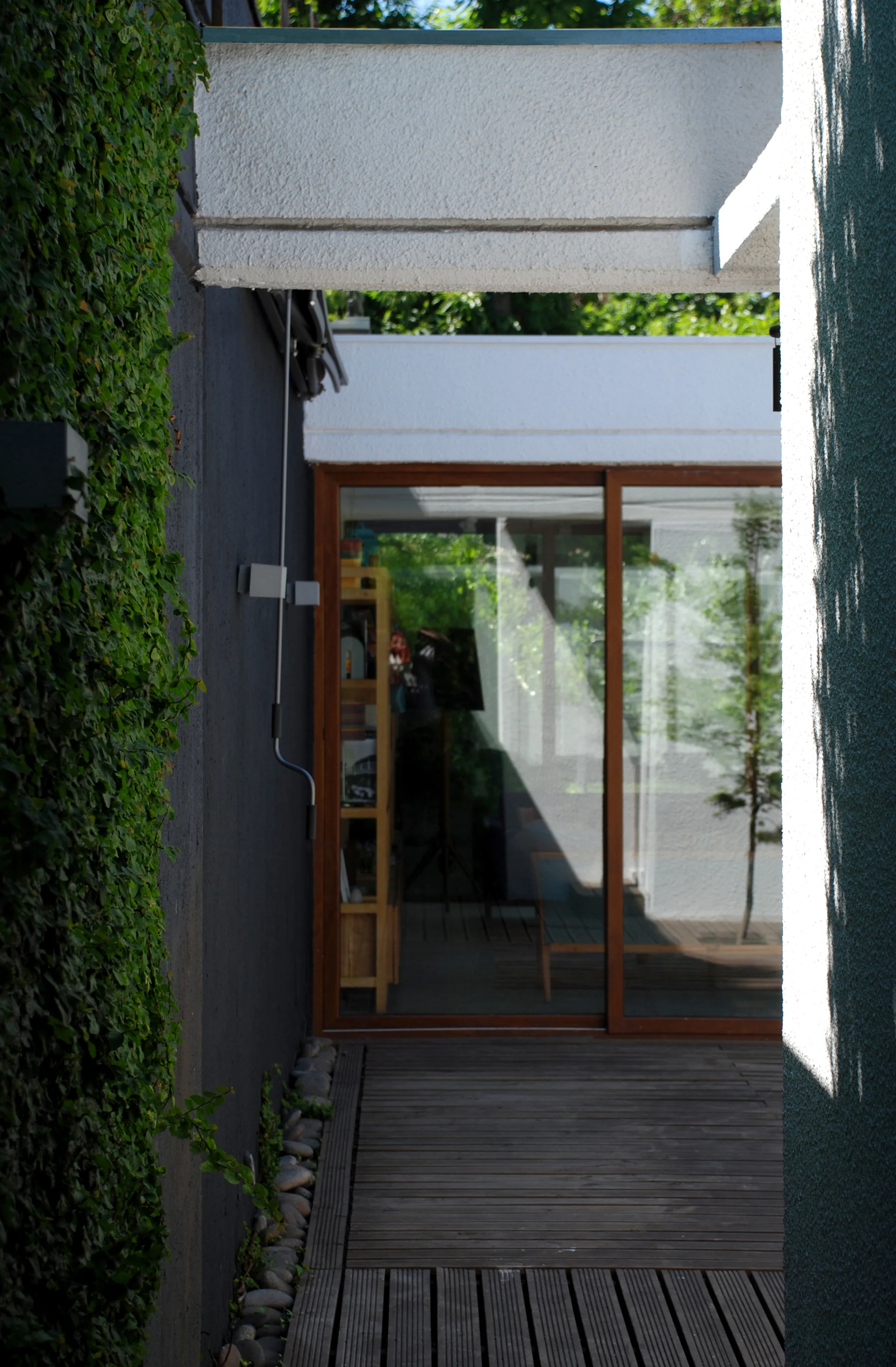 A wooden deck patio features a dark grey stucco wall covered in climbing green vines next to a sliding glass door leading into the home.
