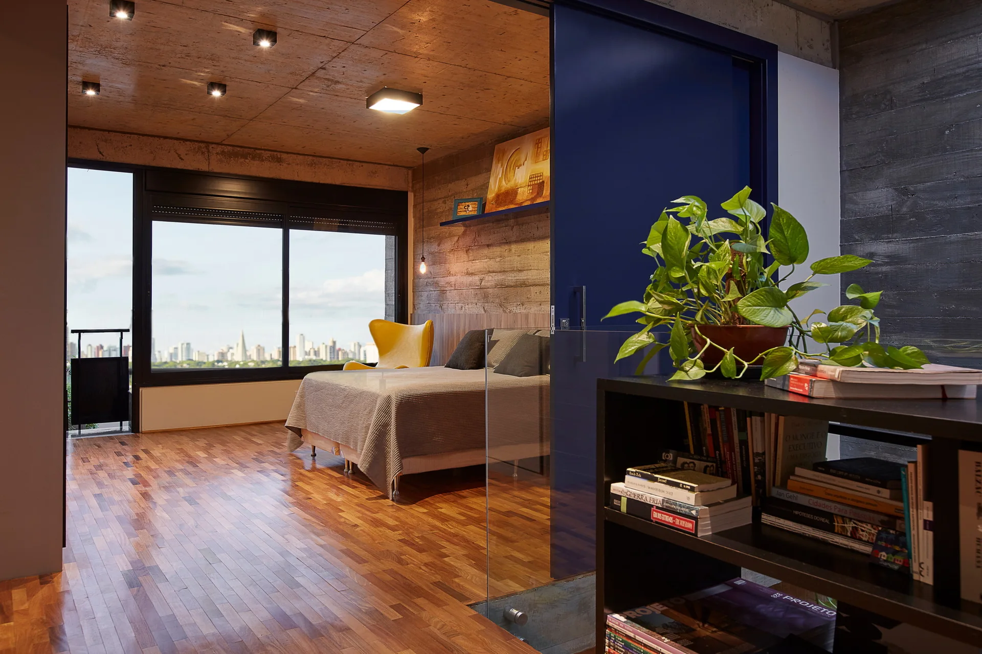 An industrial bedroom featuring an exposed concrete ceiling, parquet wood floors, a bed, a yellow accent chair, and a deep blue sliding door.