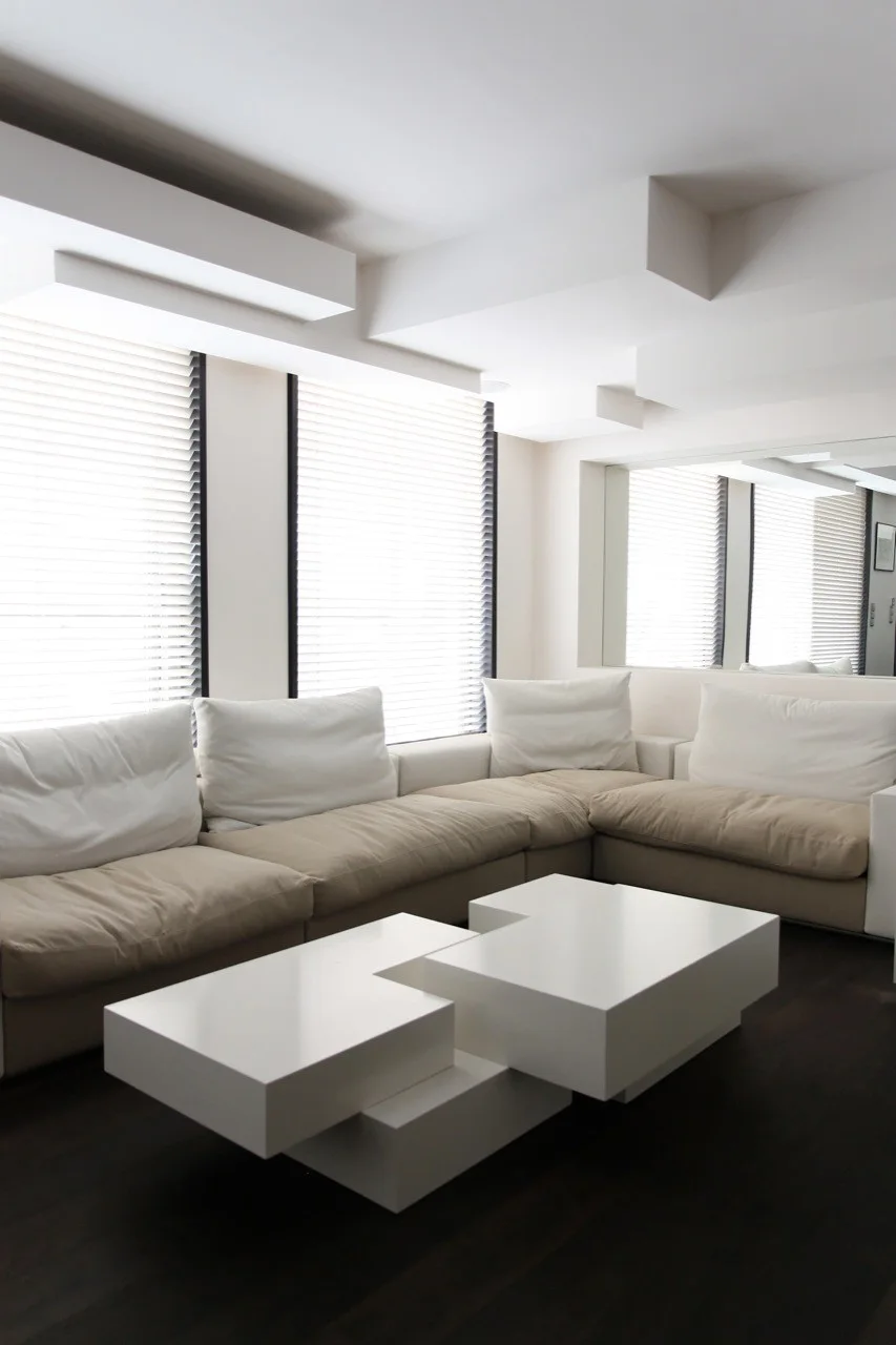 A minimalist living room features a tan sectional sofa paired with a glossy white tiered geometric coffee table and dark wood floors.