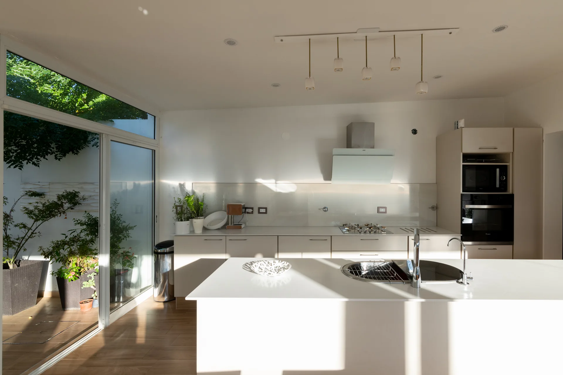 A minimalist white kitchen features a central island with a sink, flat-panel cabinets, and large sliding glass doors opening to a sunlit patio.