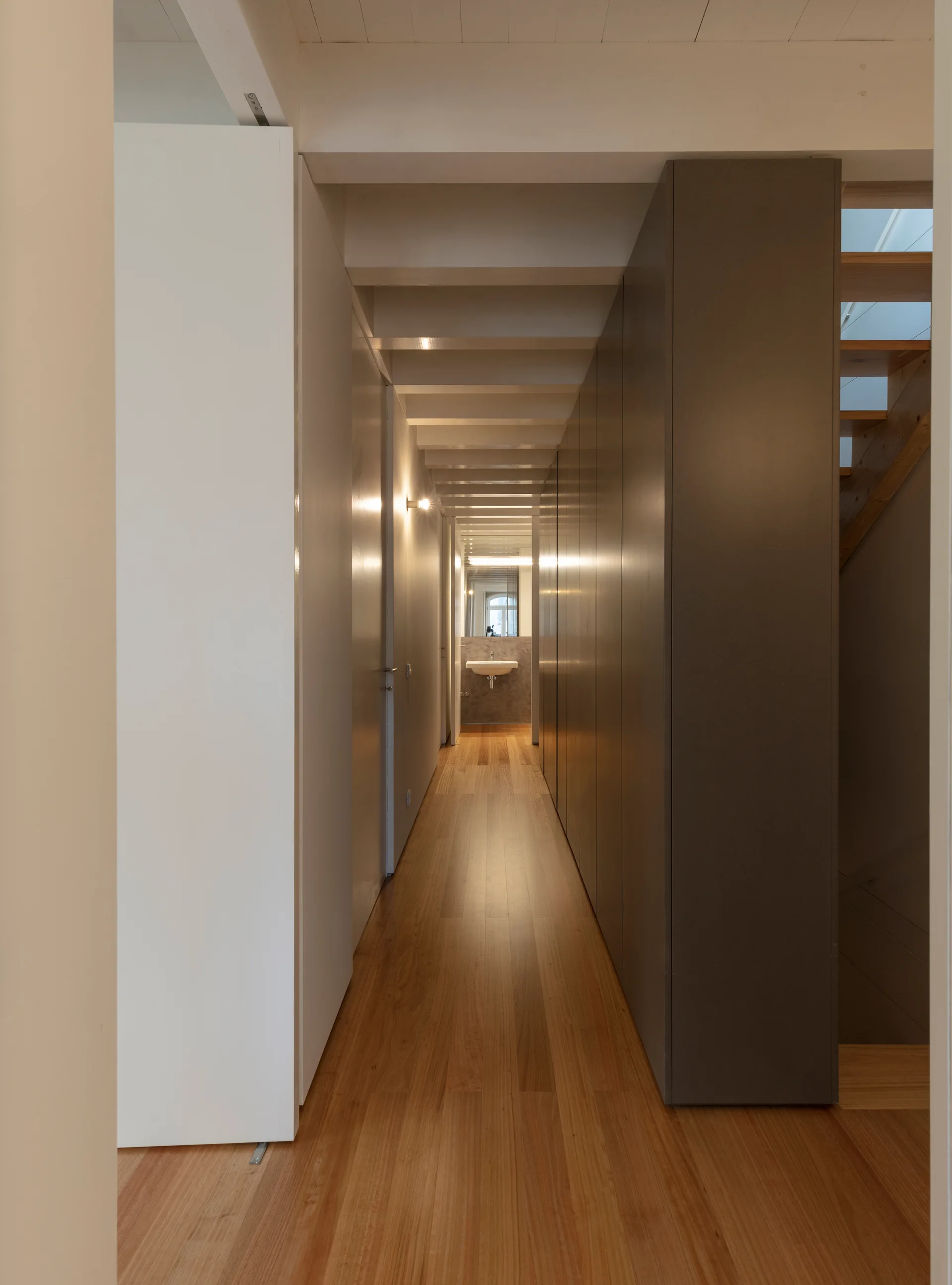 A long residential corridor features warm wood floors, white exposed ceiling beams, and a wall of tall grey built-in cabinets leading to a distant sink.
