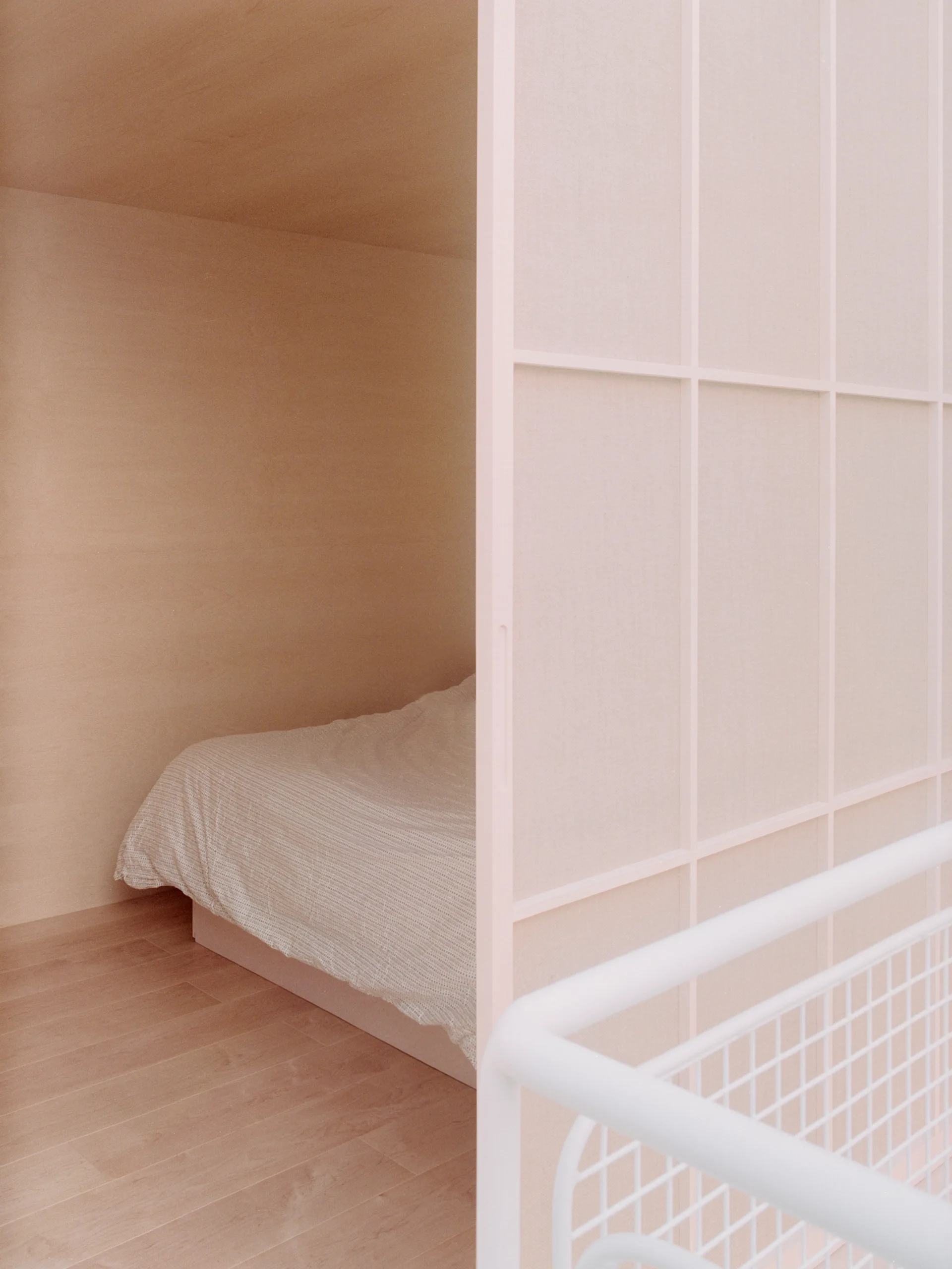 A bed rests inside a light wood-paneled alcove next to a large, pale pink grid partition screen in a minimalist room.