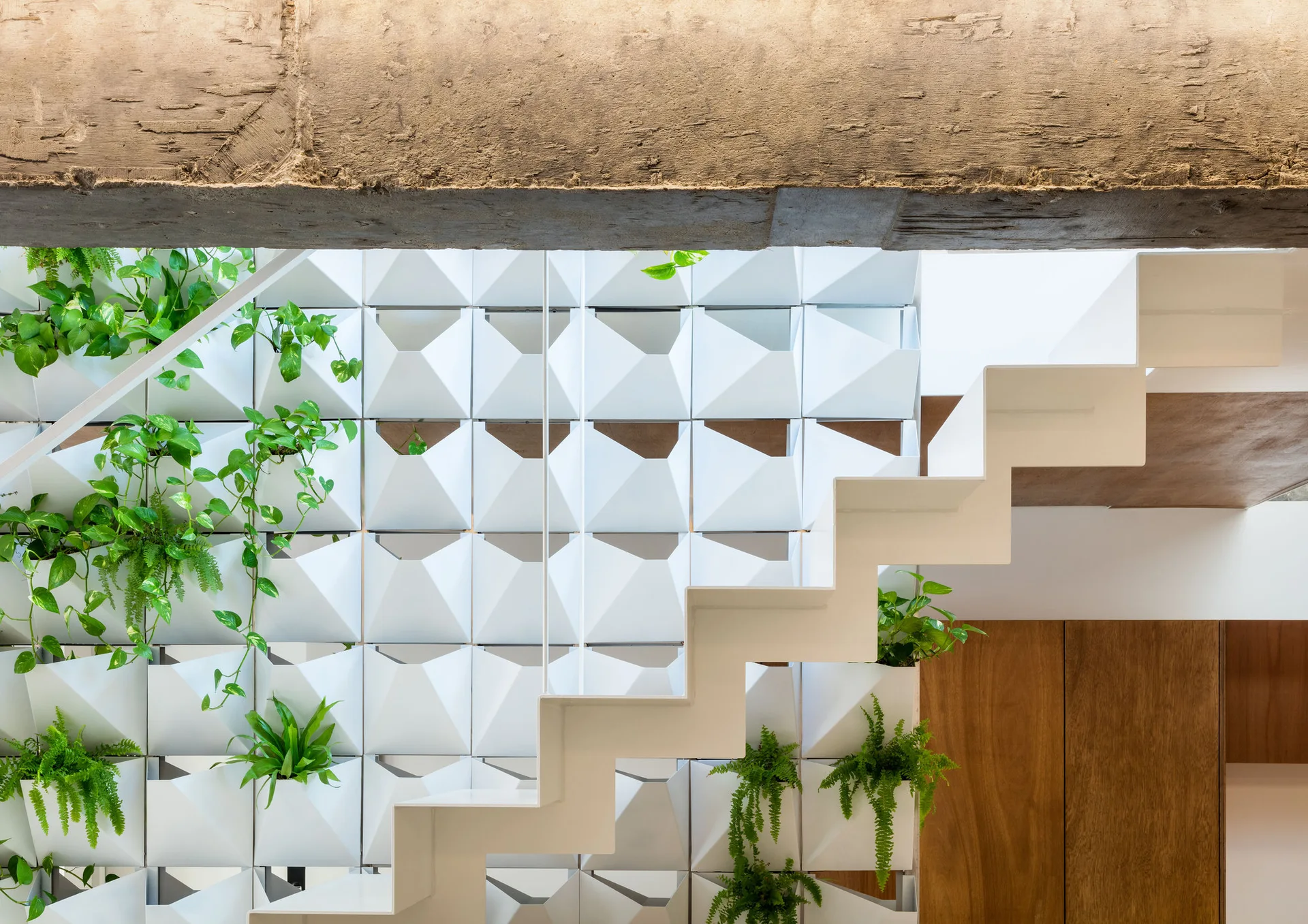 A white zigzag staircase ascends past a geometric modular wall featuring built-in planters with green leafy plants under a weathered beam.