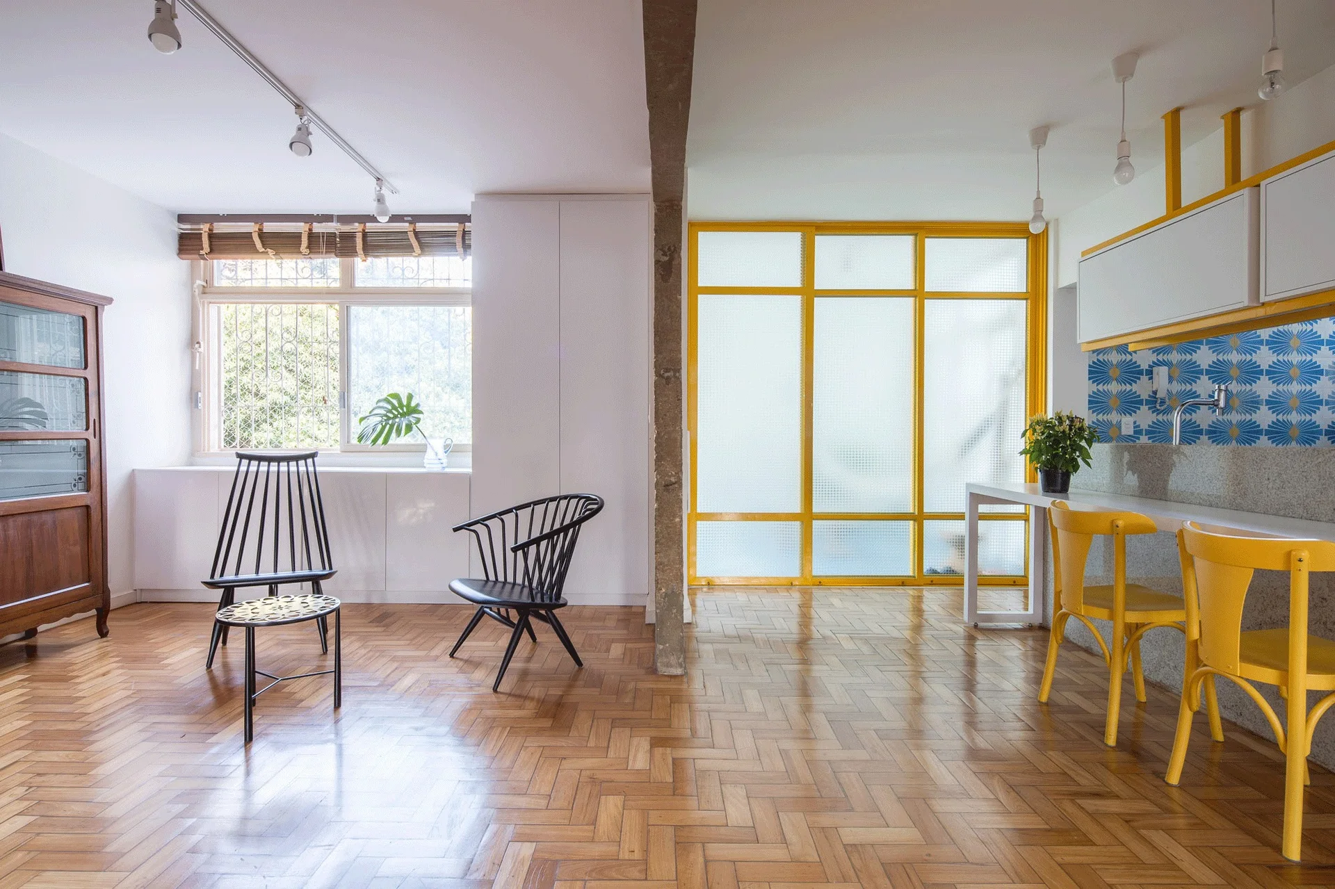 An open plan kitchen and living room featuring herringbone wood floors, yellow stools, and a yellow-framed glass partition wall.