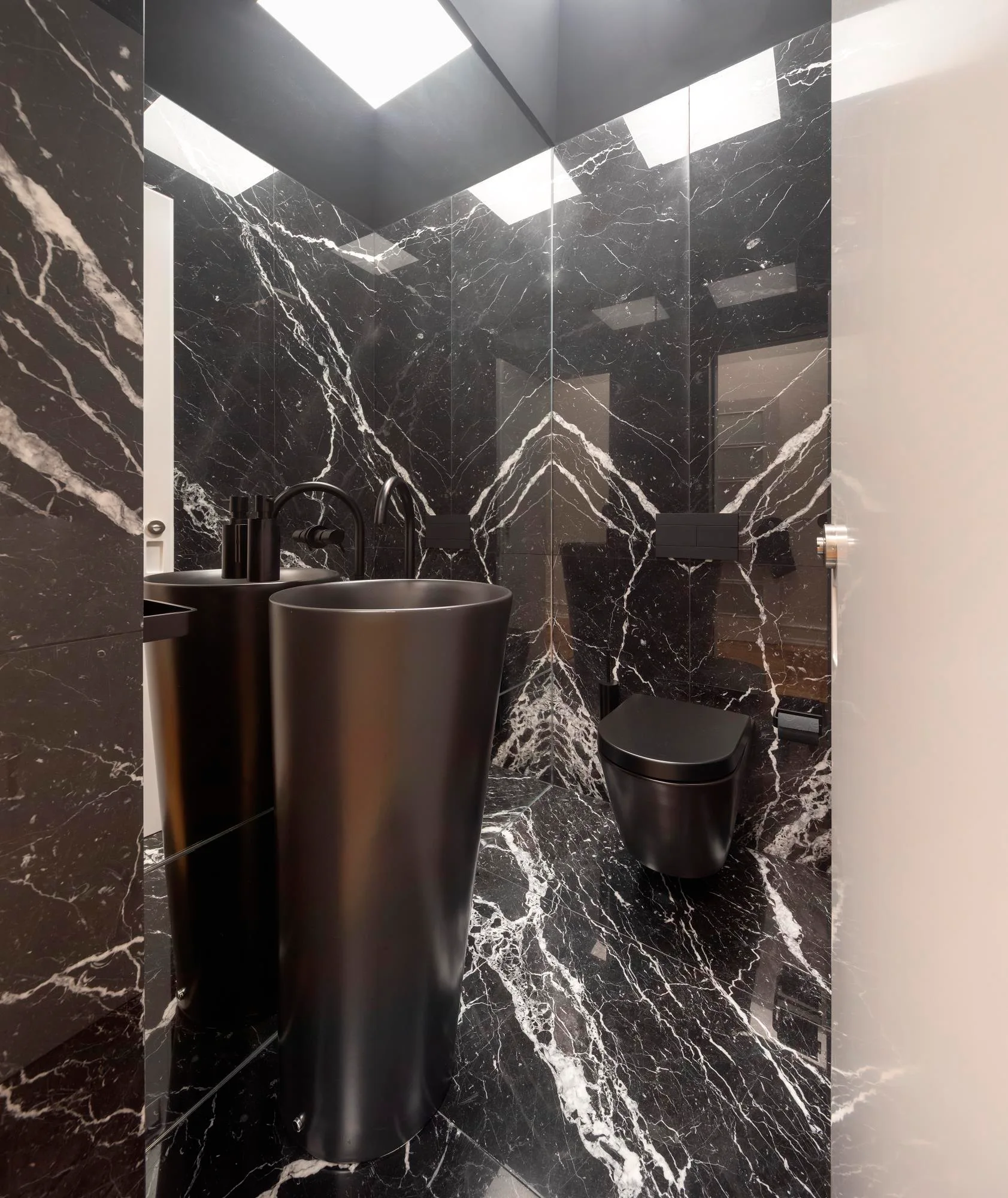 A contemporary bathroom features polished black marble walls with bold white veining, a matte black pedestal sink, and a wall-mounted toilet.