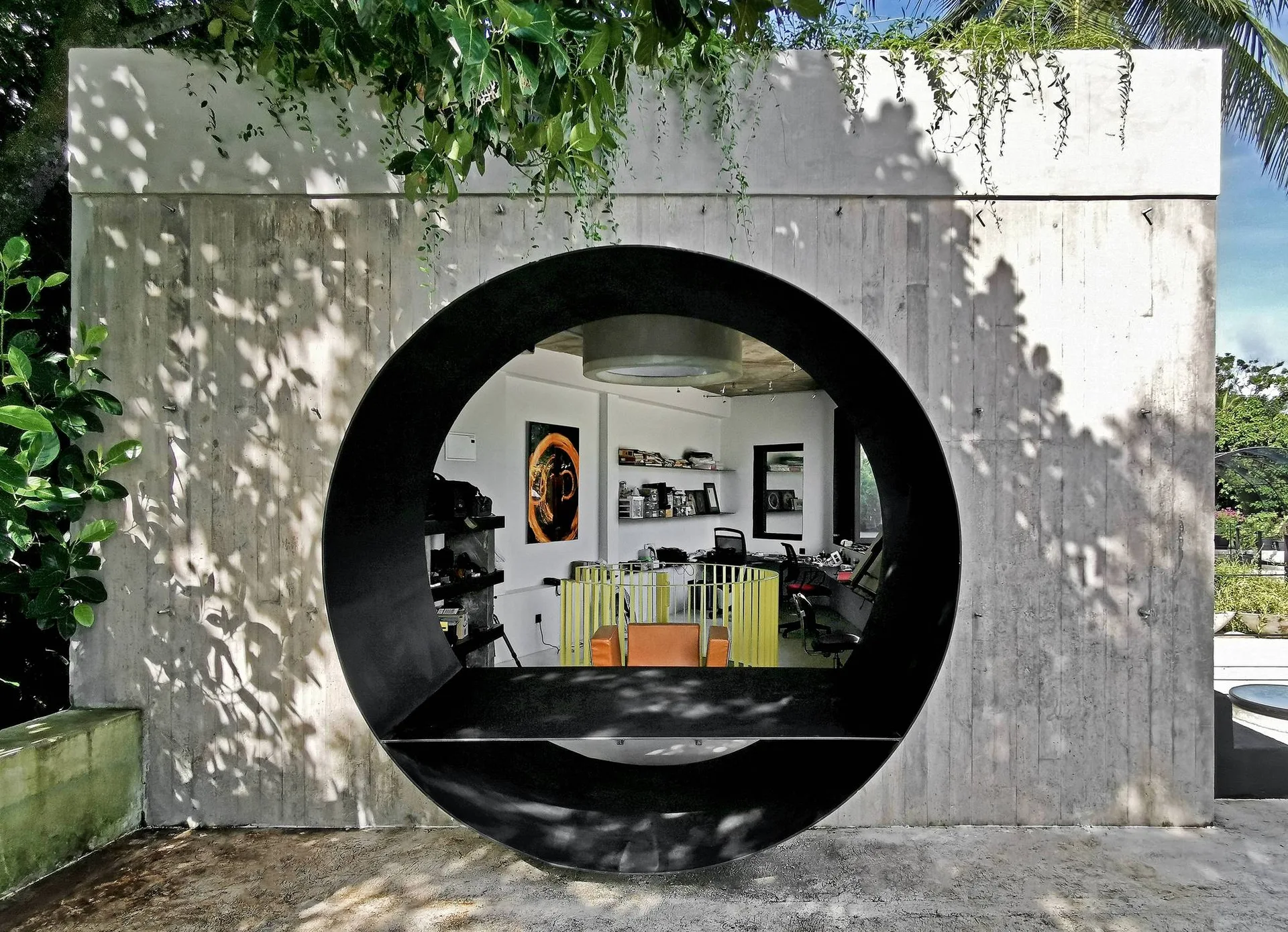 A large circular black-framed window set in a textured concrete wall reveals a modern home office with desks and colorful seating inside.