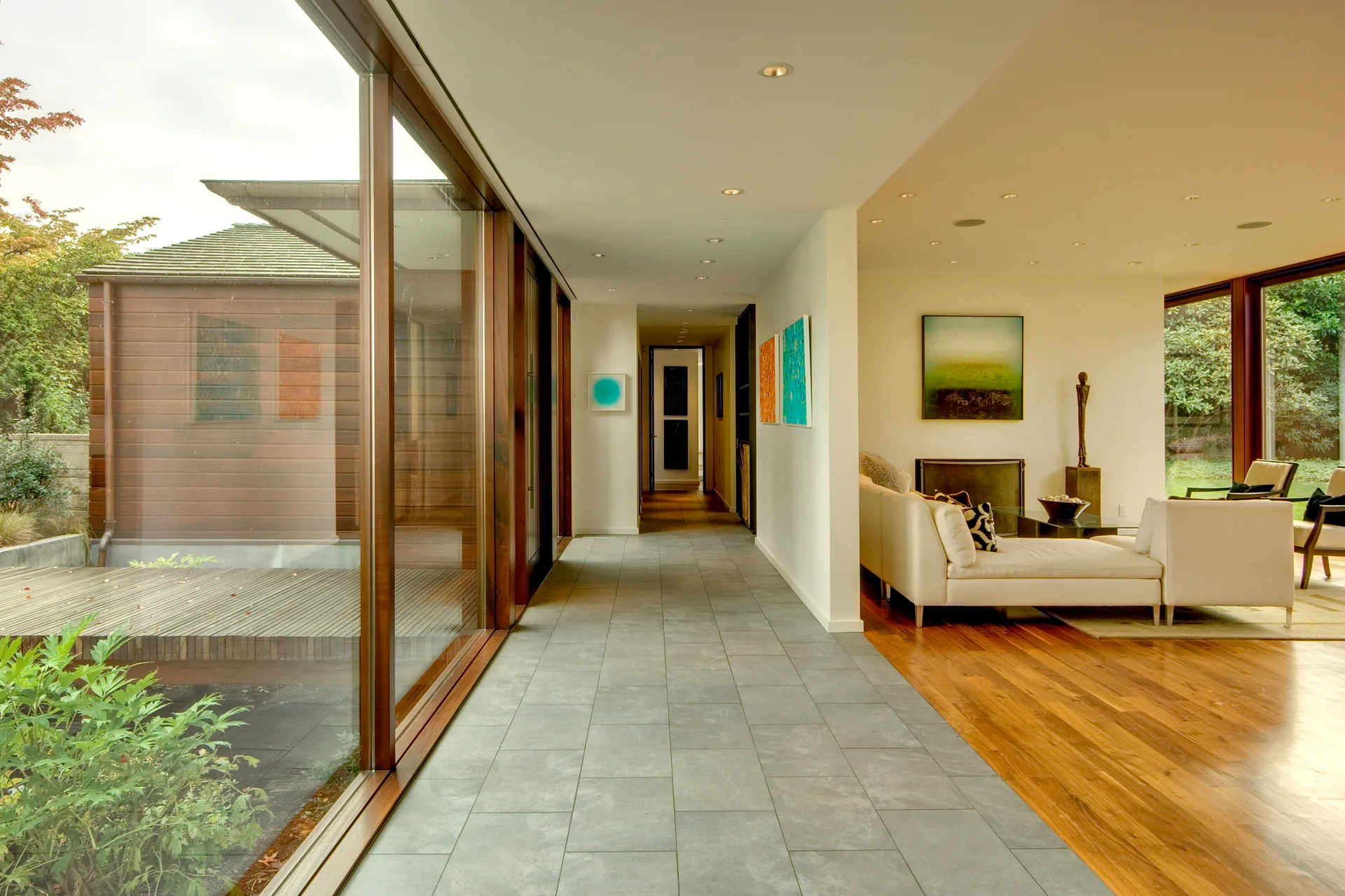 A long hallway with grey floor tiles transitions into a living room with warm hardwood floors and large wood-framed windows.
