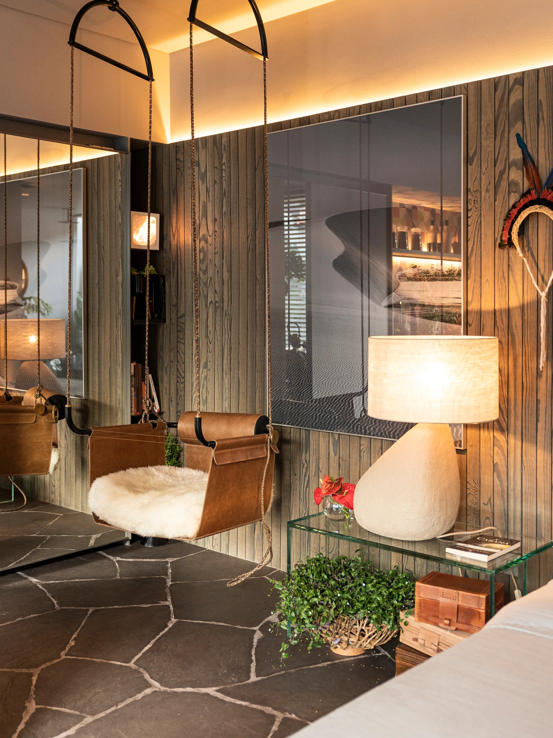 An indoor lounge featuring a leather hanging swing chair, vertical wood wall paneling, a stone floor, and a glowing table lamp.