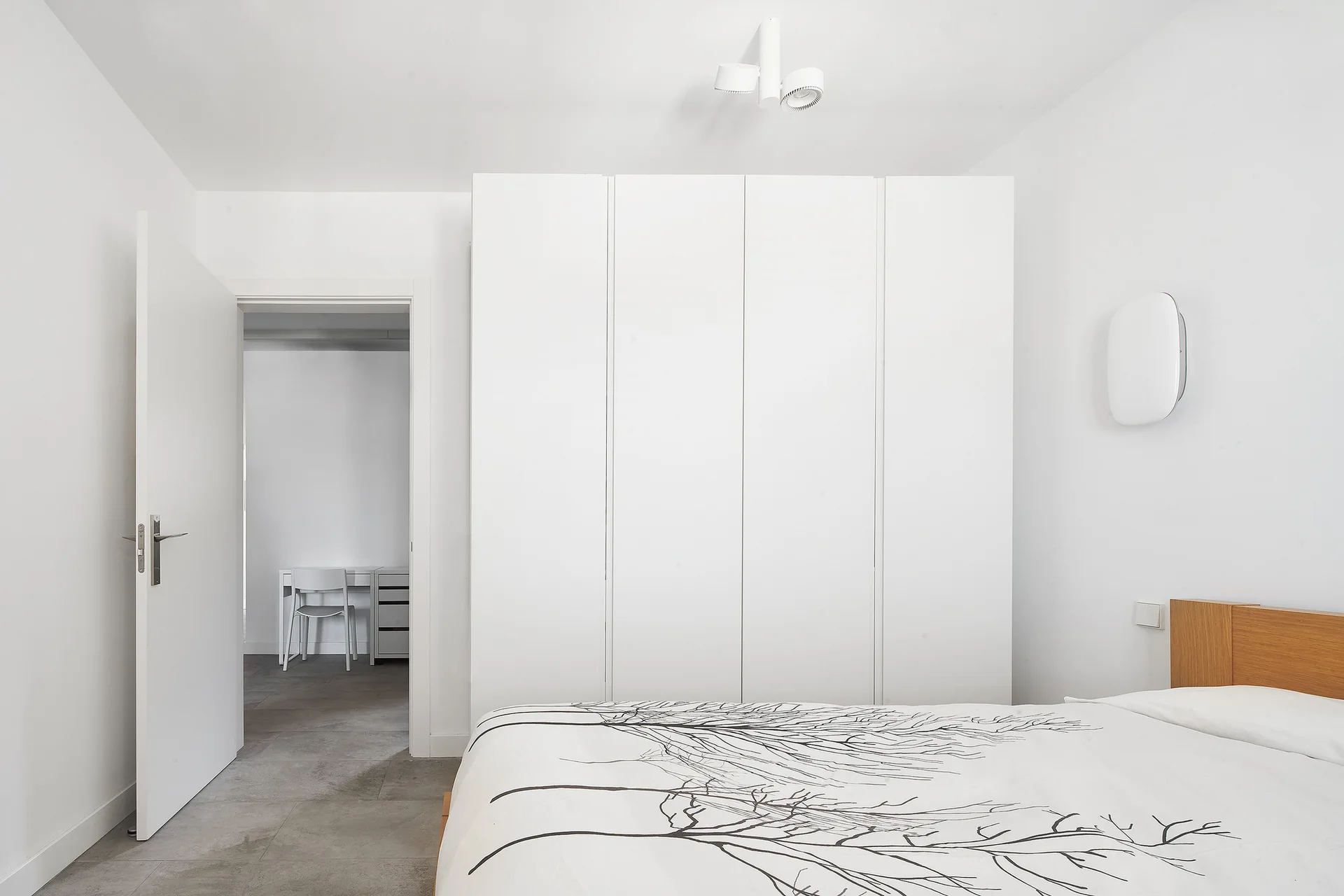 A minimalist white bedroom features a tall built-in wardrobe, a bed with a tree-patterned duvet, and an open door showing a small home office.