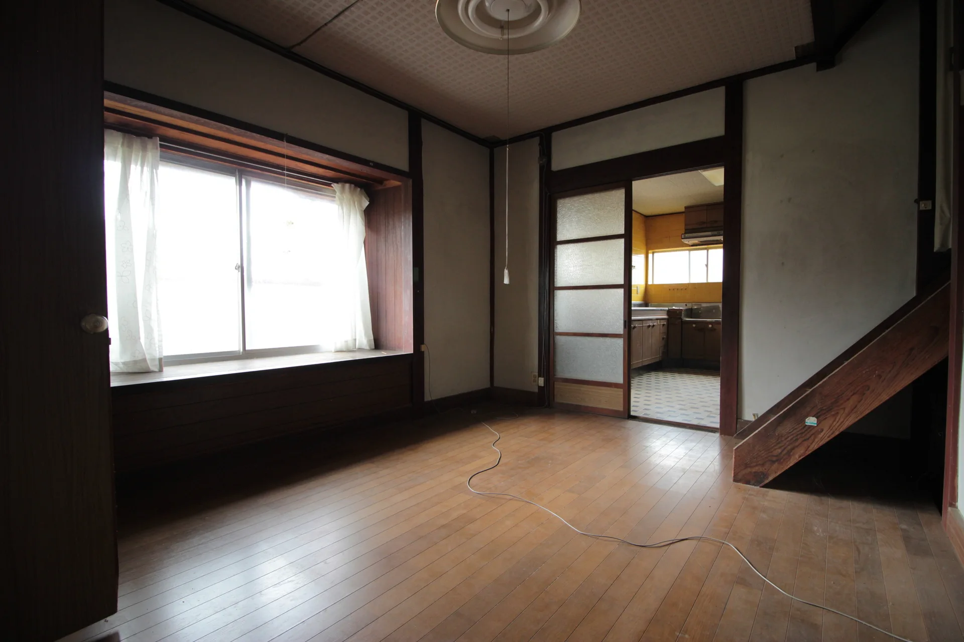 An empty Japanese-style room features warm wood flooring, a deep wooden window frame with white curtains, and a sliding glass partition door.