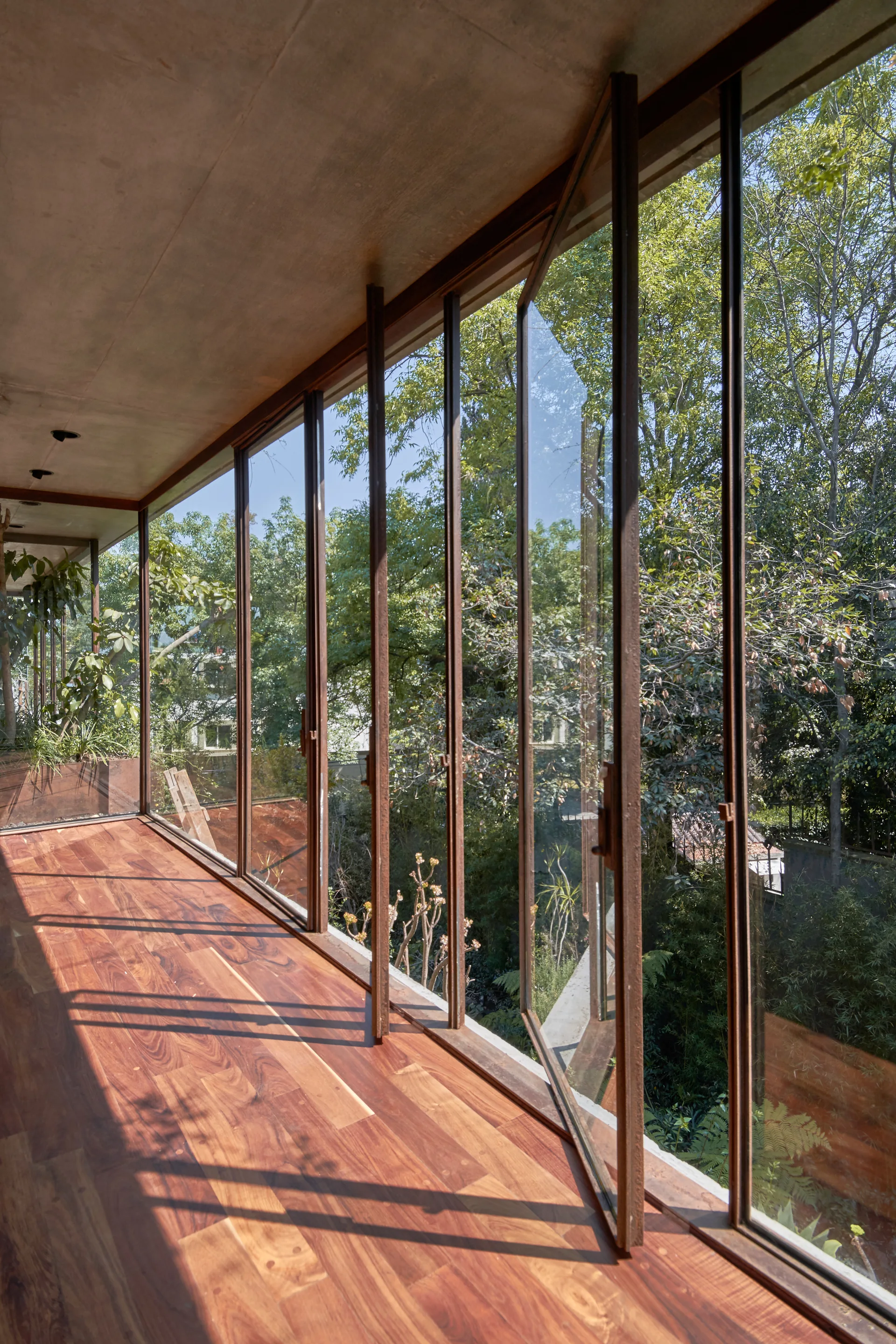 A minimalist corridor with rich wood flooring, a concrete ceiling, and floor-to-ceiling pivoting glass doors that open to a garden.