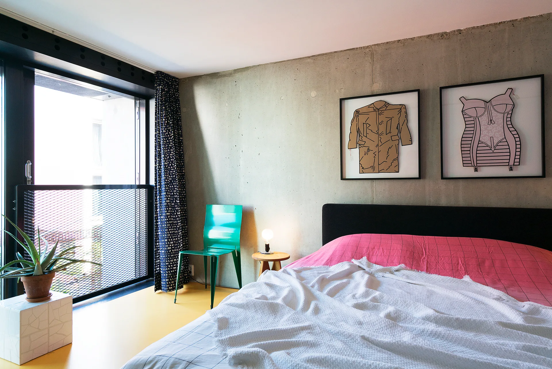 A modern bedroom featuring a bed with pink bedding, an exposed concrete wall with two framed art pieces, a green metal chair, and a bright yellow floor.