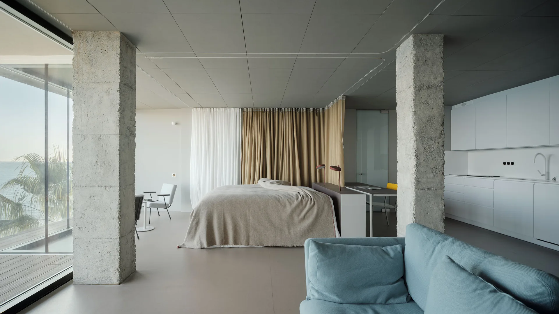 A minimalist studio apartment featuring raw concrete columns, a bed partitioned by floor-to-ceiling curtains, a light blue sofa, and a white kitchen.