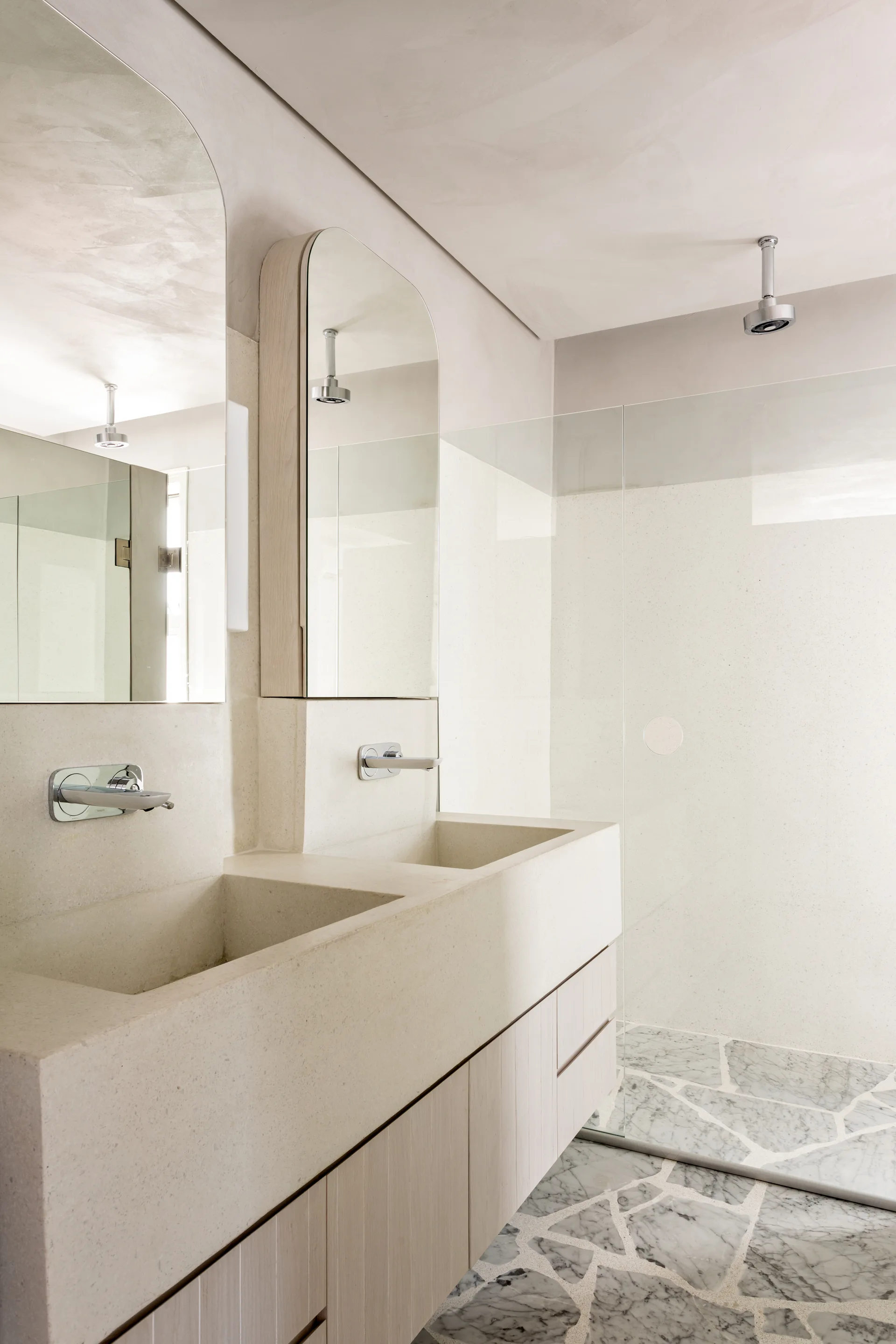 A minimalist double bathroom vanity features an integrated beige stone sink, arched wooden mirrors, wall-mounted faucets, and irregular marble floor tile.